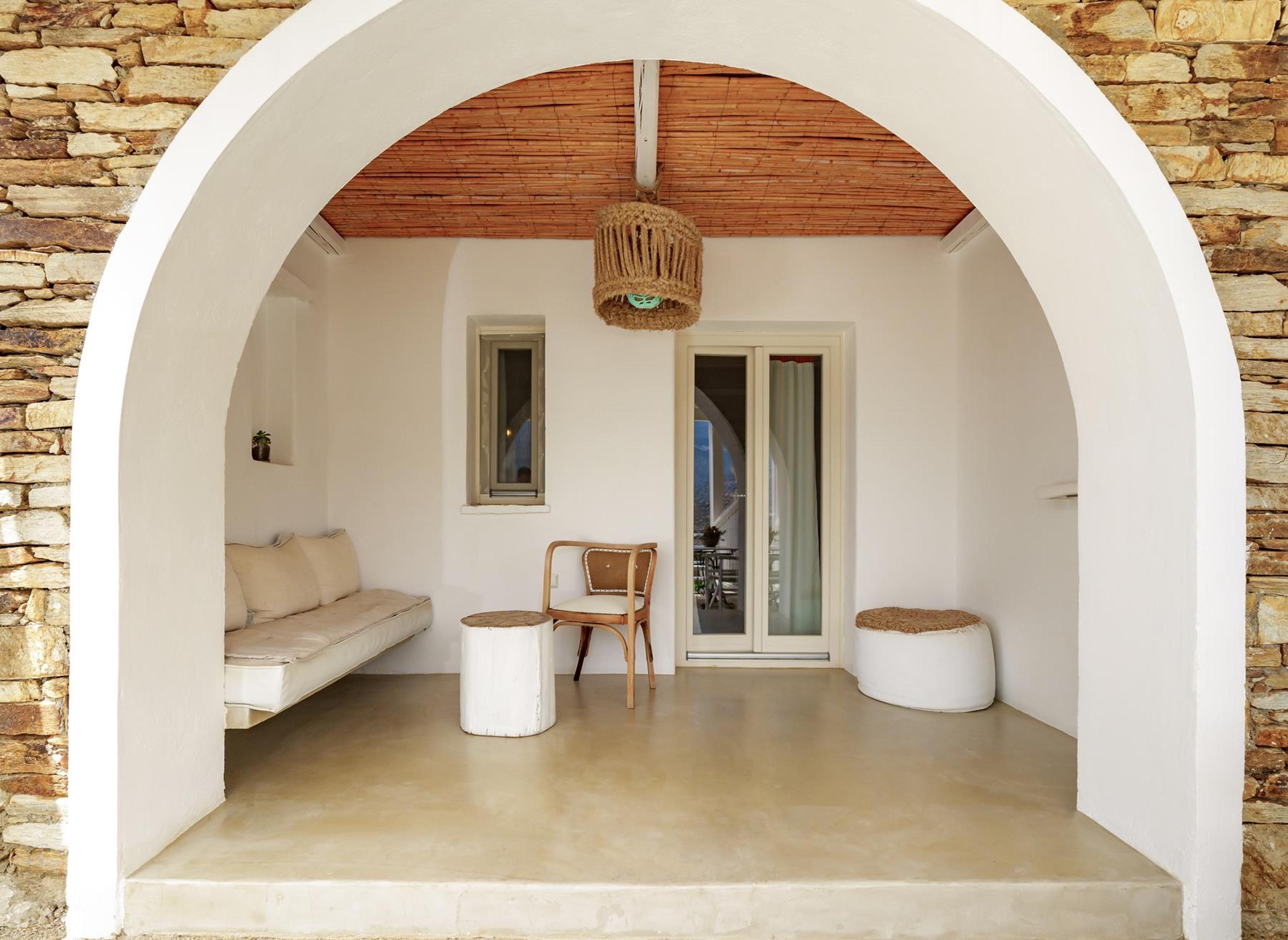 An arched doorway in a stone wall, opening onto a small terrace with a cushioned daybed, a wicker chair and a white stool.