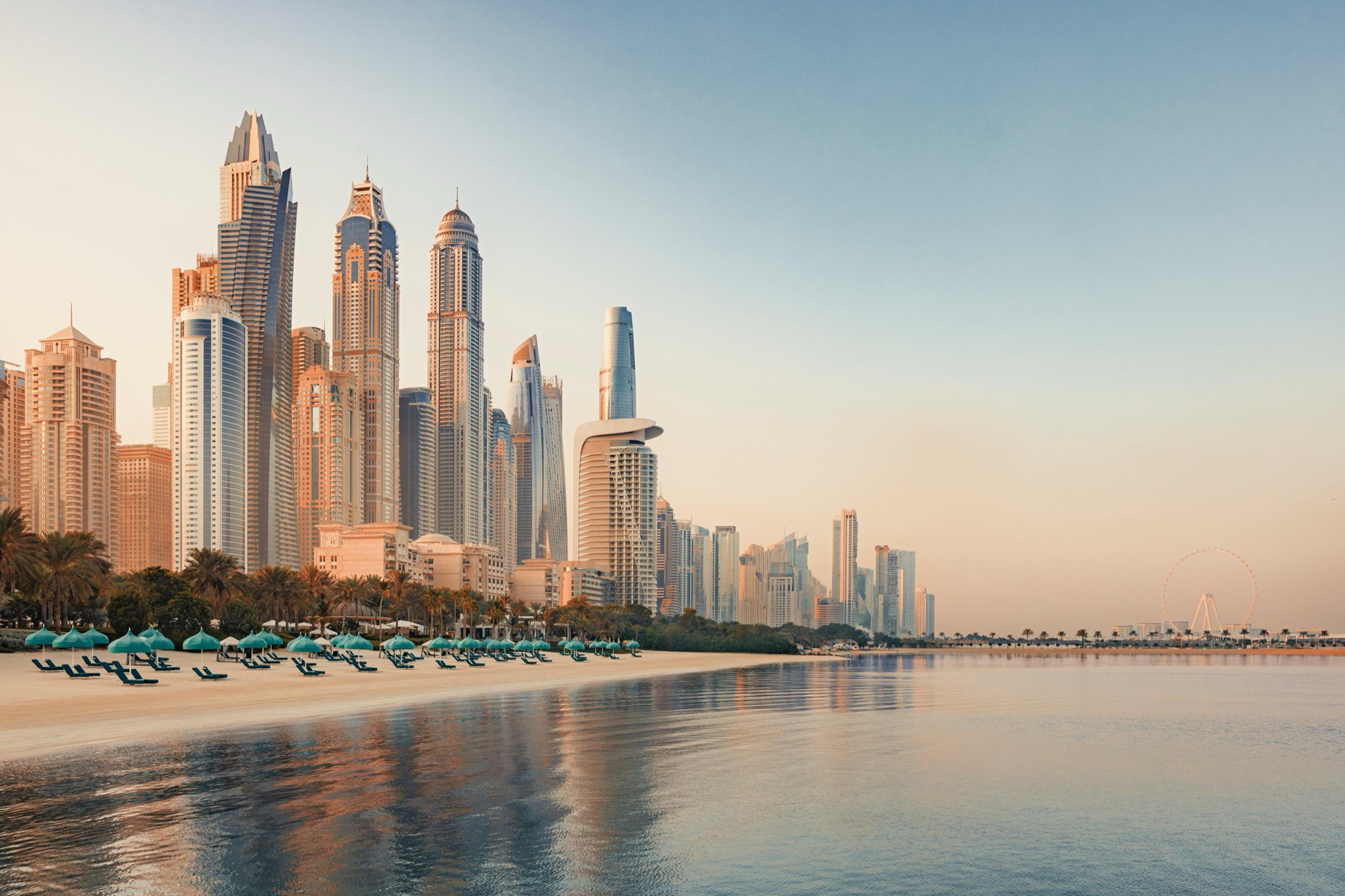 A row of lit beachfront umbrellas sits below a cluster of high-rise towers reflected in calm water at dawn.