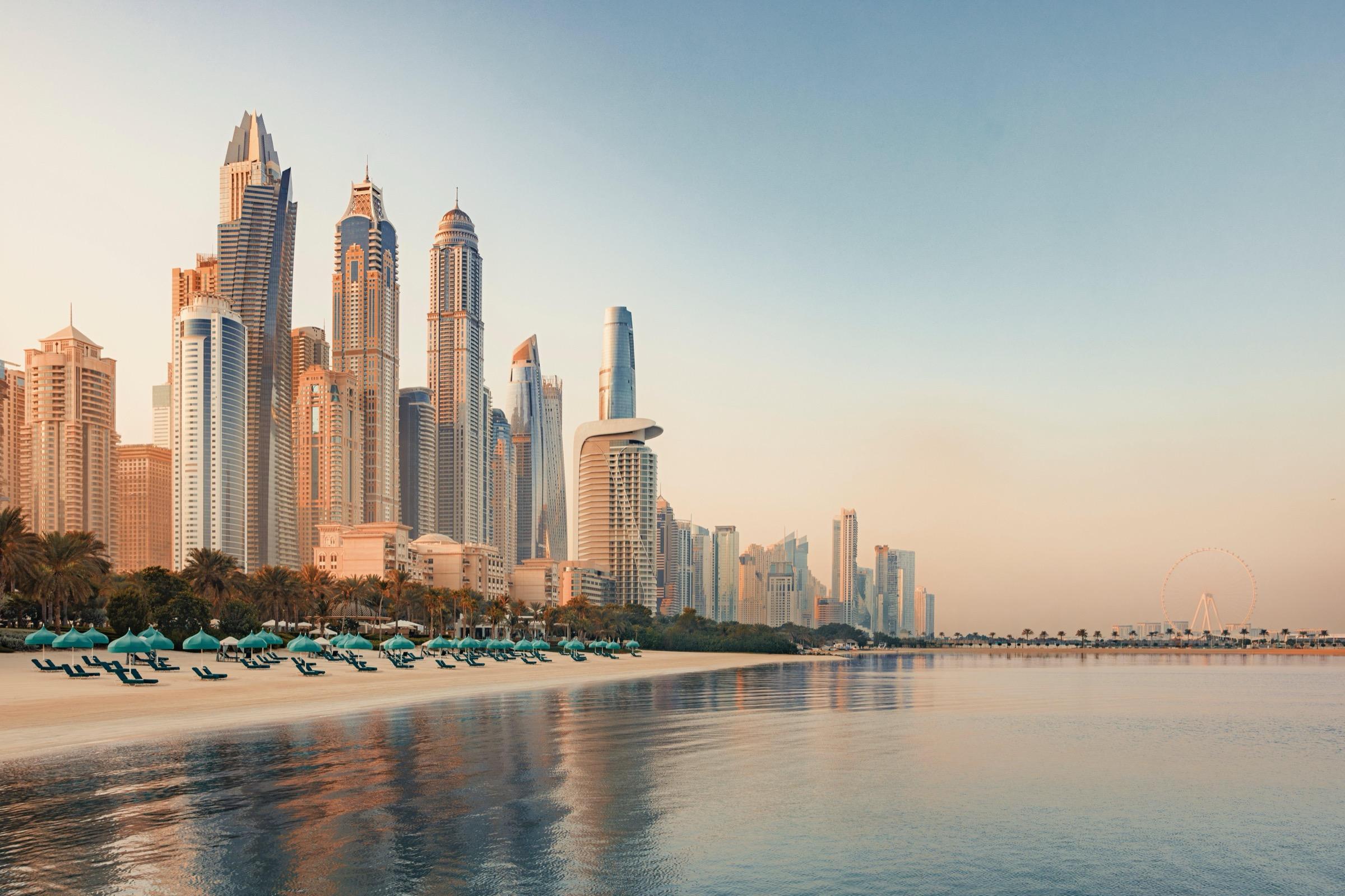 A row of lit beachfront umbrellas sits below a cluster of high-rise towers reflected in calm water at dawn.