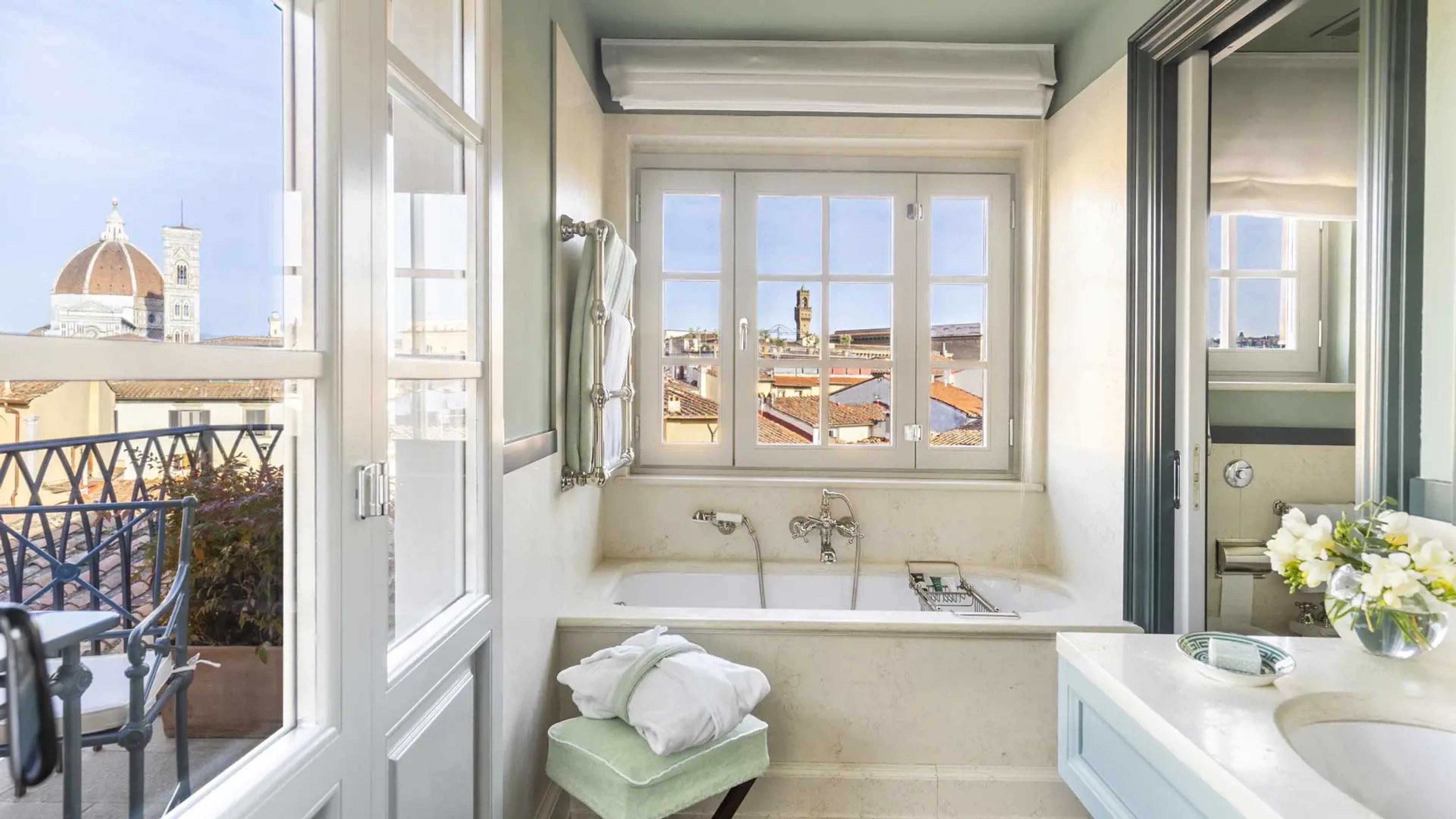 A freestanding bathtub beneath a window framing the Duomo's dome and bell tower, with white towels on a rail and a chair beside the tub, seen through an open door from a terrace.