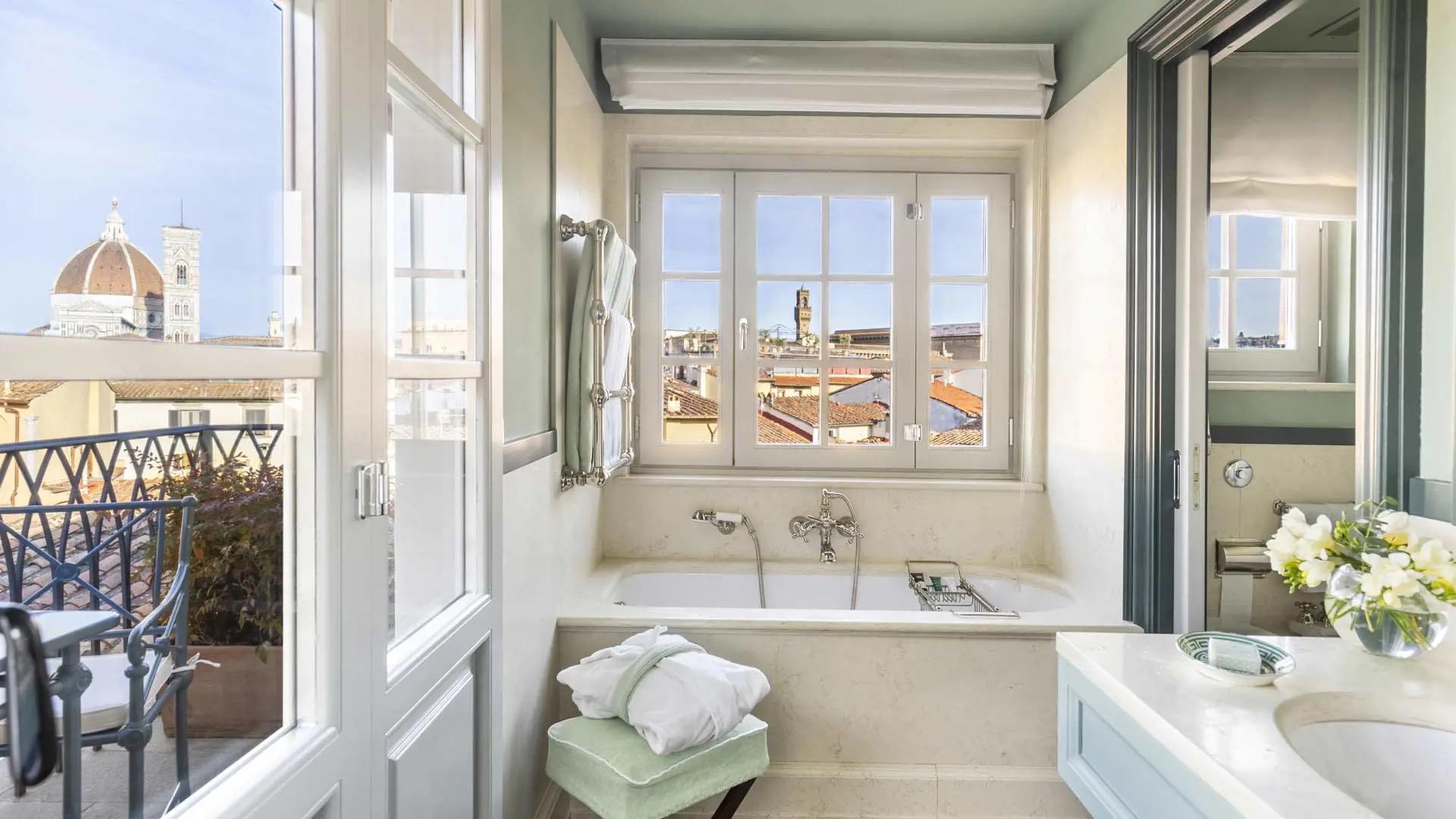 A freestanding bathtub beneath a window framing the Duomo's dome and bell tower, with white towels on a rail and a chair beside the tub, seen through an open door from a terrace.