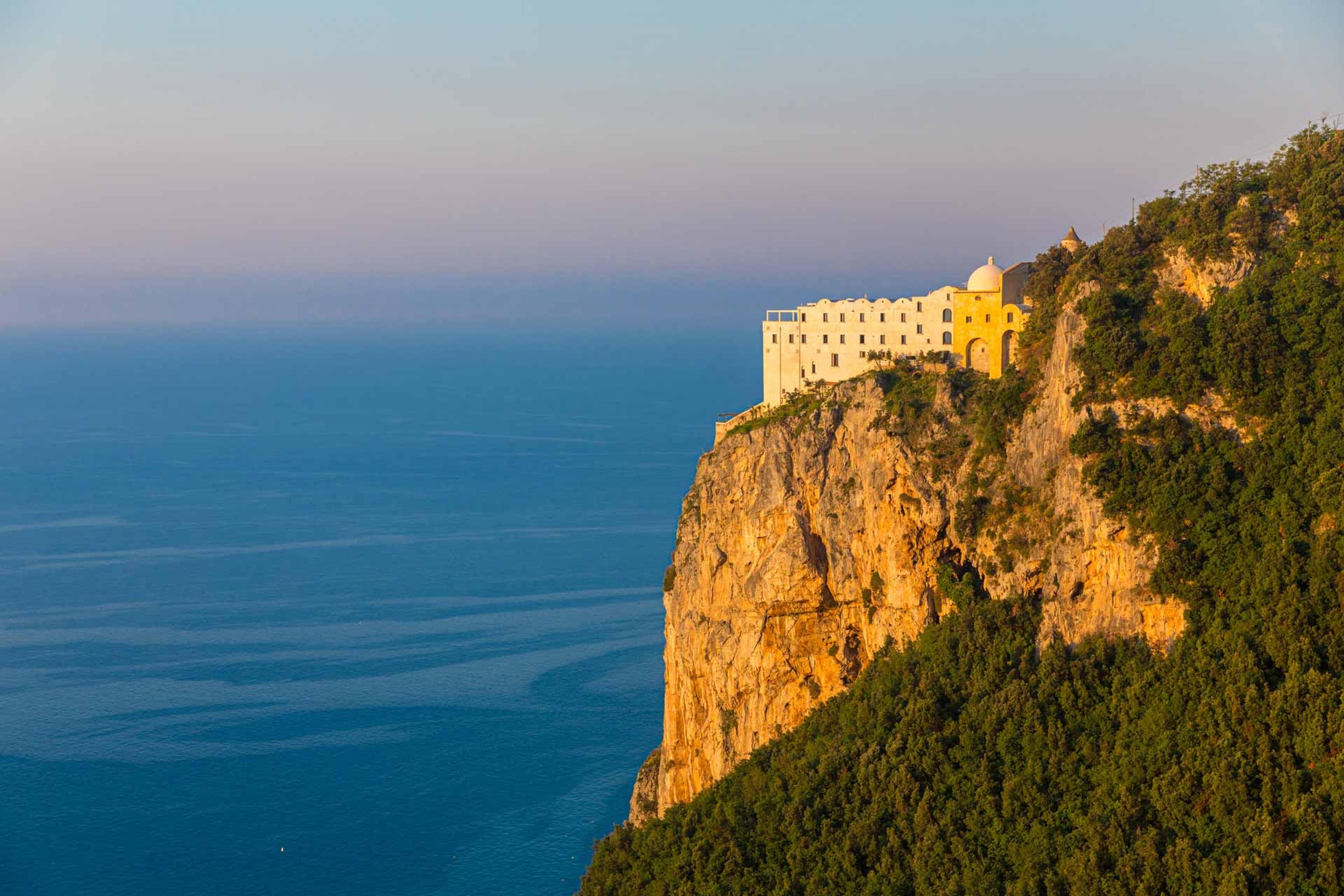 the monastery's pale buildings lit gold at sunset on a cliff above the sea, with a wooded hillside to the right