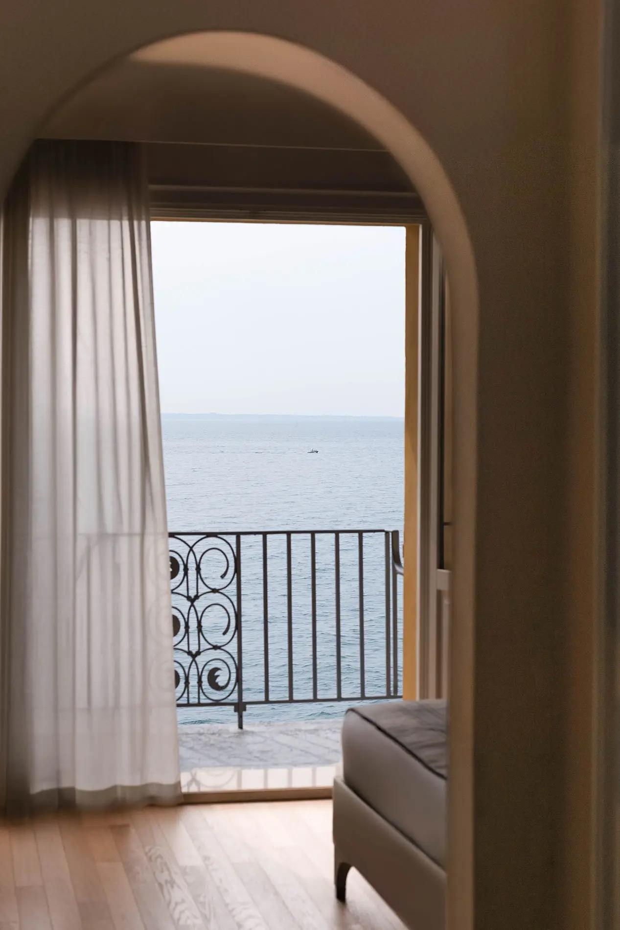 A curtained archway leads to a wrought iron balcony railing and open lake water, the foot of a bed in the foreground.