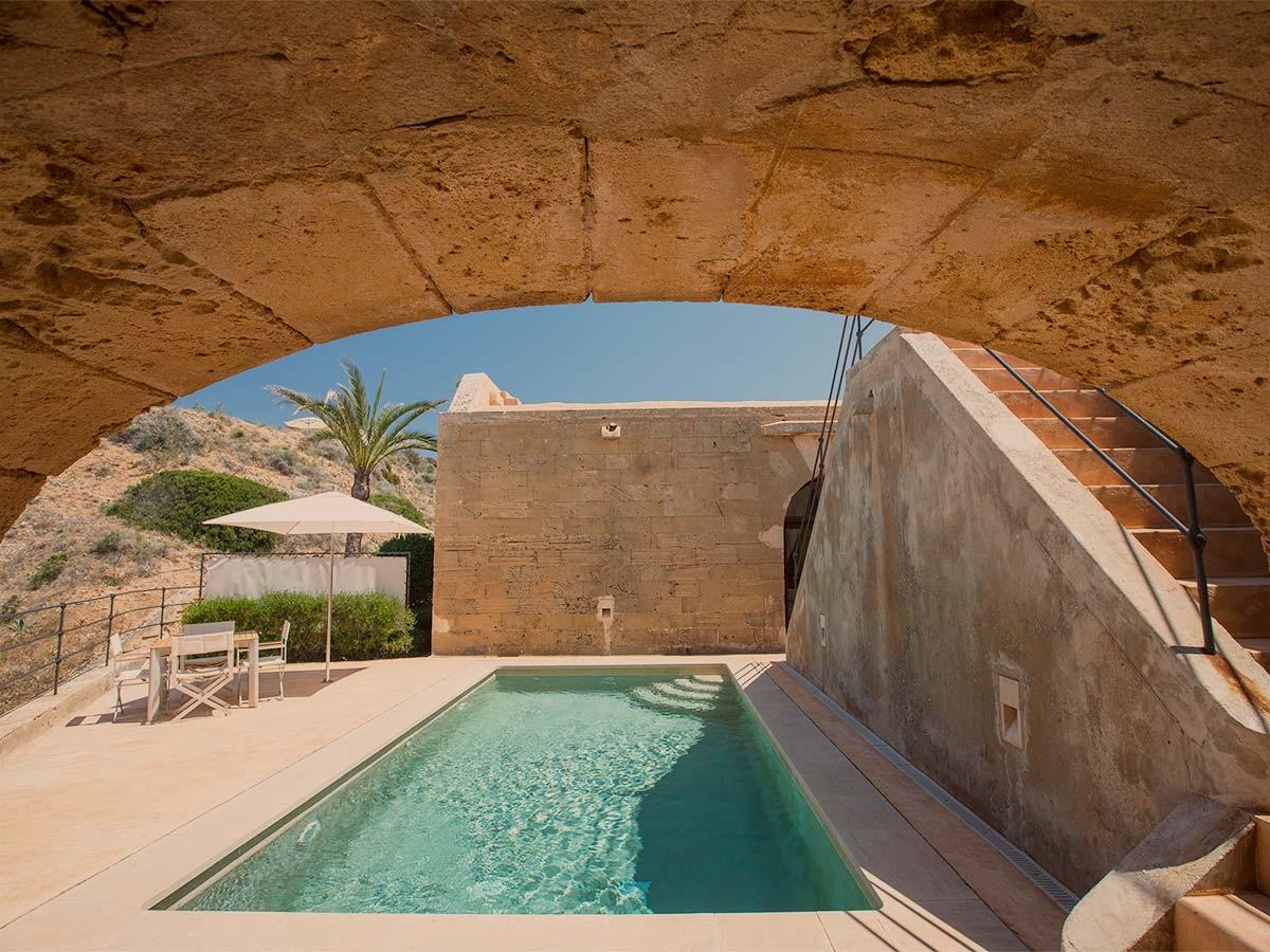 A swimming pool seen through a stone archway, with a terrace, chairs, and a parasol beside it and hills visible in the distance.