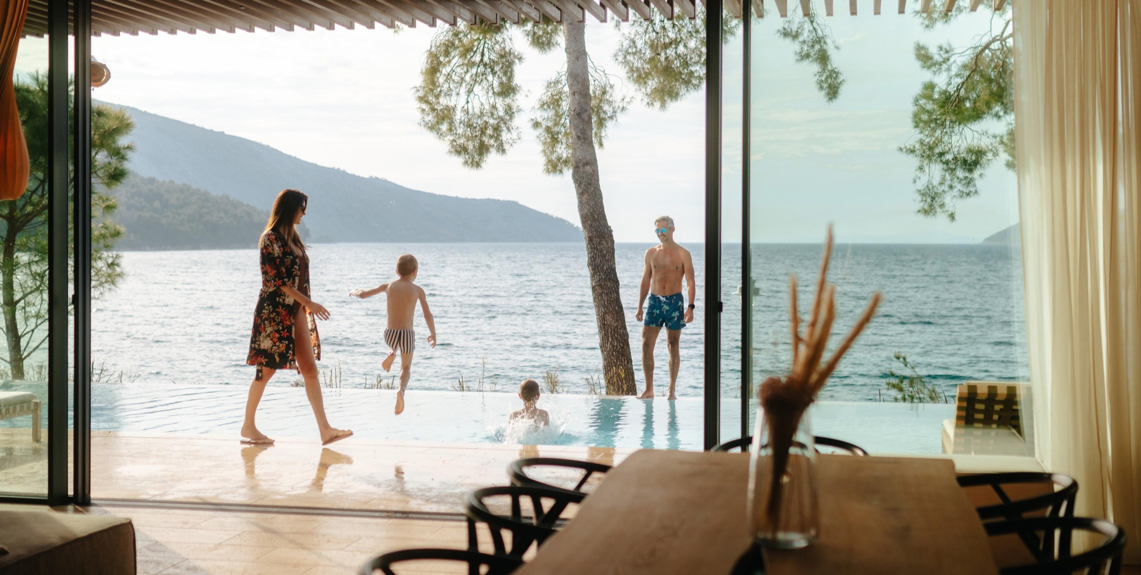 A family by an infinity pool, a boy jumping in and a small child in the water, with the sea and a hill behind, seen through a glass wall.