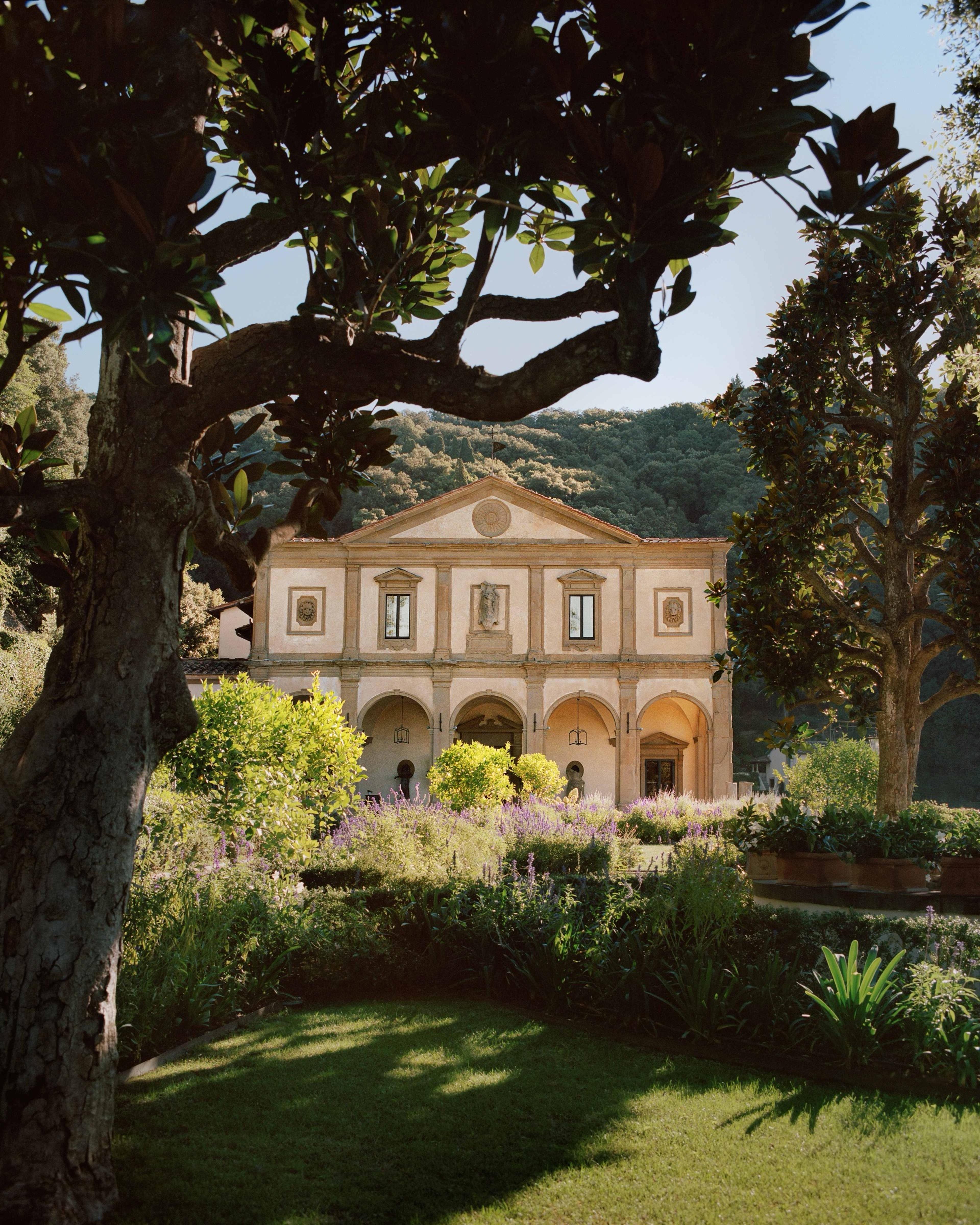 A pink stone facade with a pedimented front and arched ground-floor windows stands behind clipped garden beds, seen through the branches of a tree in the foreground, with wooded hills rising behind.