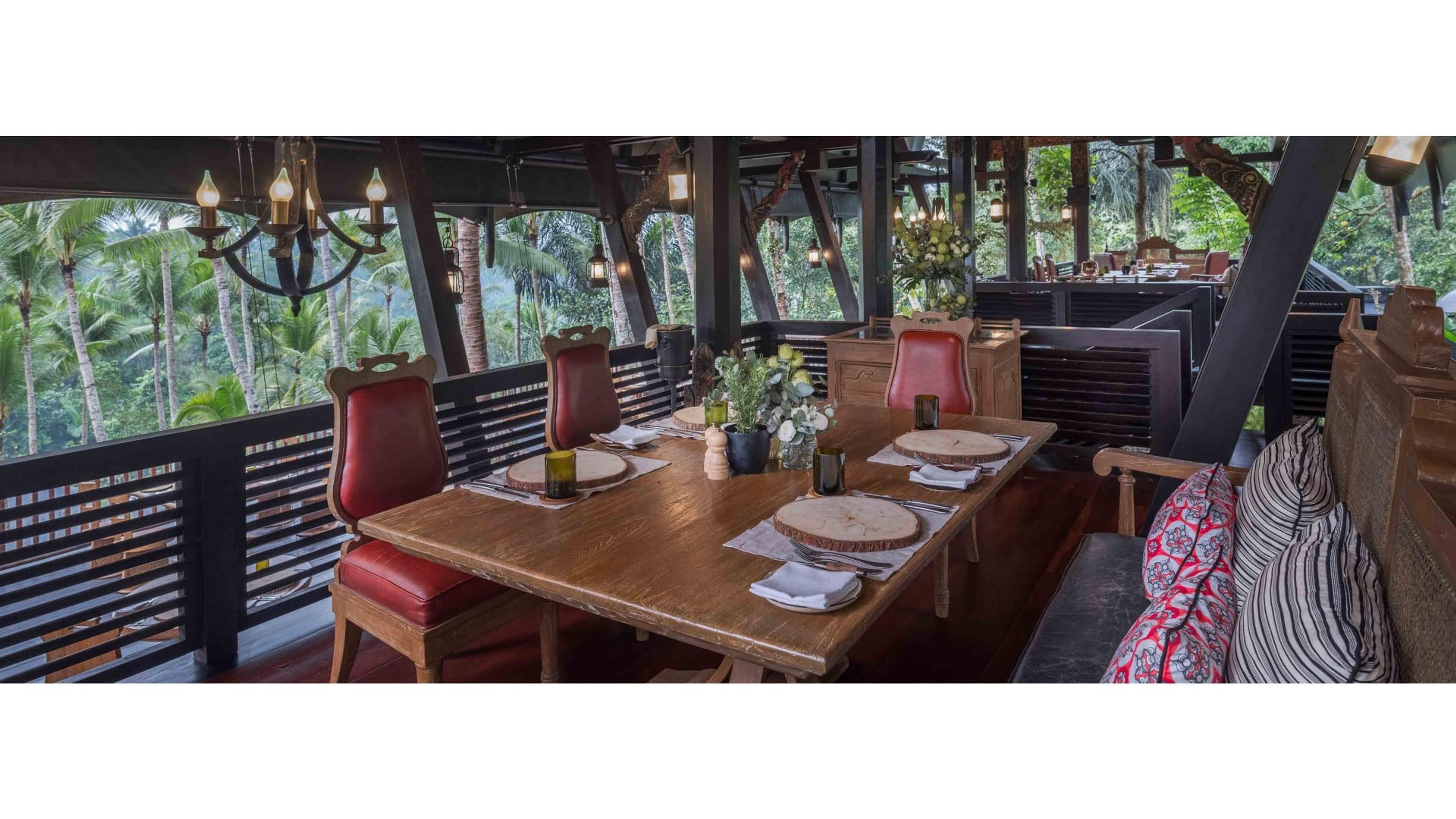 A wooden dining table set for two with charger plates and folded napkins, flanked by red leather chairs, on an open-sided terrace overlooking a forested valley, with a chandelier hanging above.