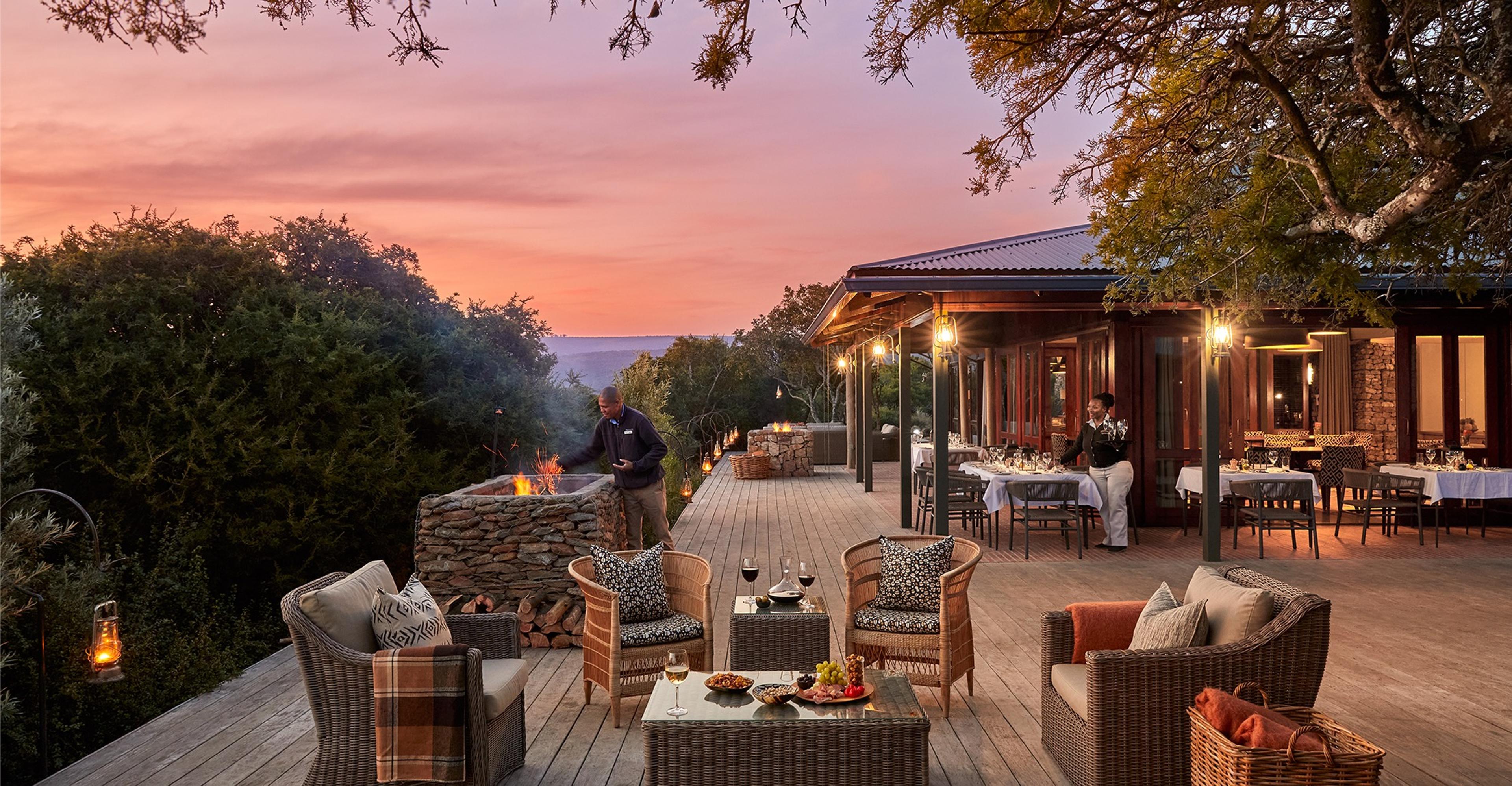 A lodge terrace at dusk with wicker armchairs around a stone fire pit, and a lit dining room with set tables under a thatched roof behind.