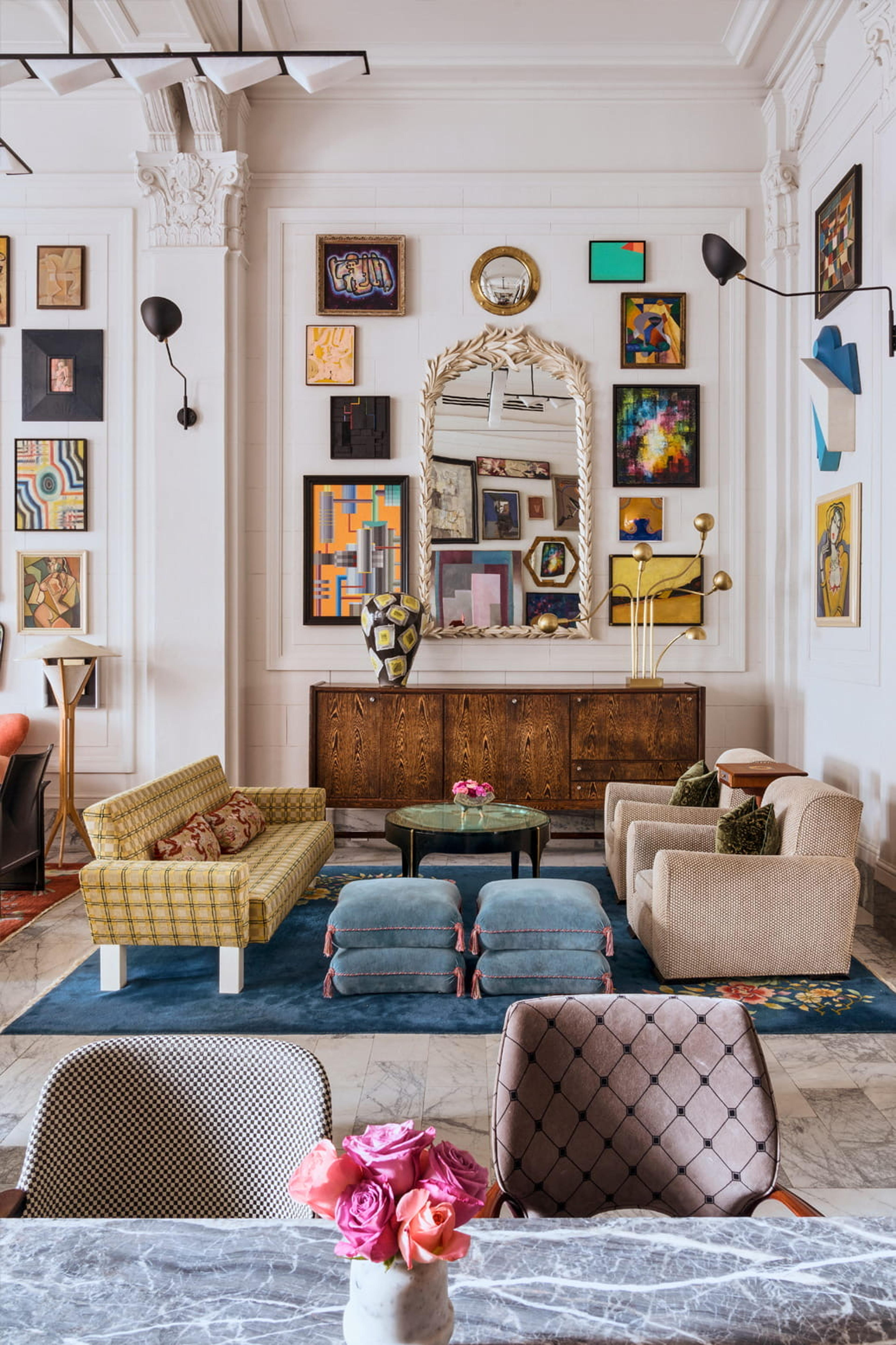 A wide lobby lounge with a dense gallery wall of framed art above patterned sofas and armchairs, a carved mirror over a wooden sideboard, and a marble floor with a blue rug.