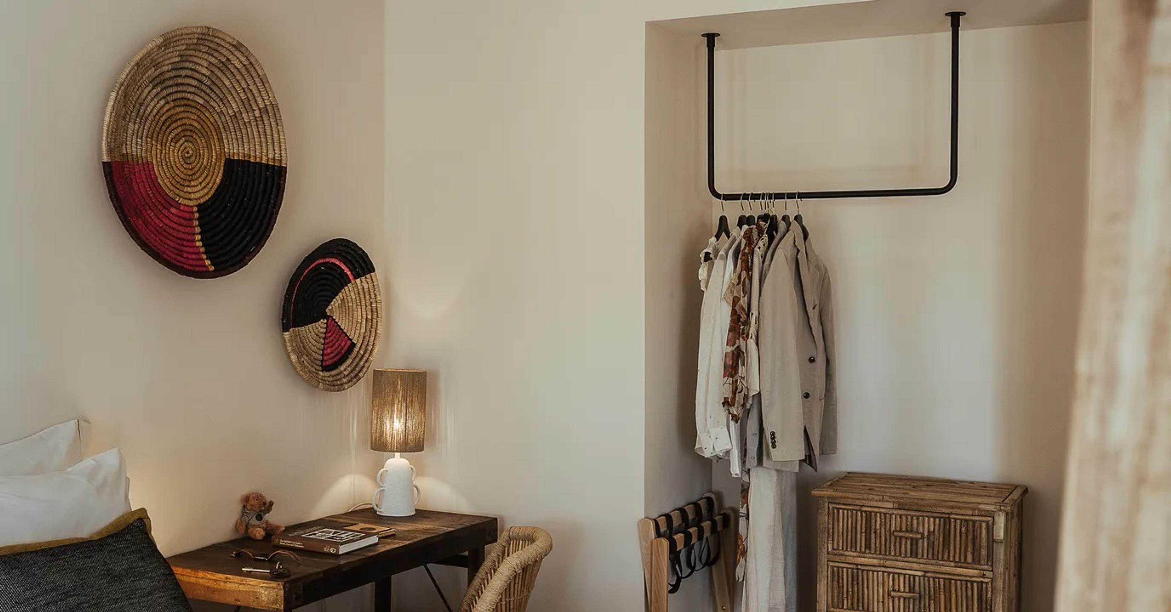 A bedroom corner with two round woven wall baskets, a wooden desk holding books and sunglasses, a cane chair, and a clothes rail hung with garments beside a rattan chest of drawers.