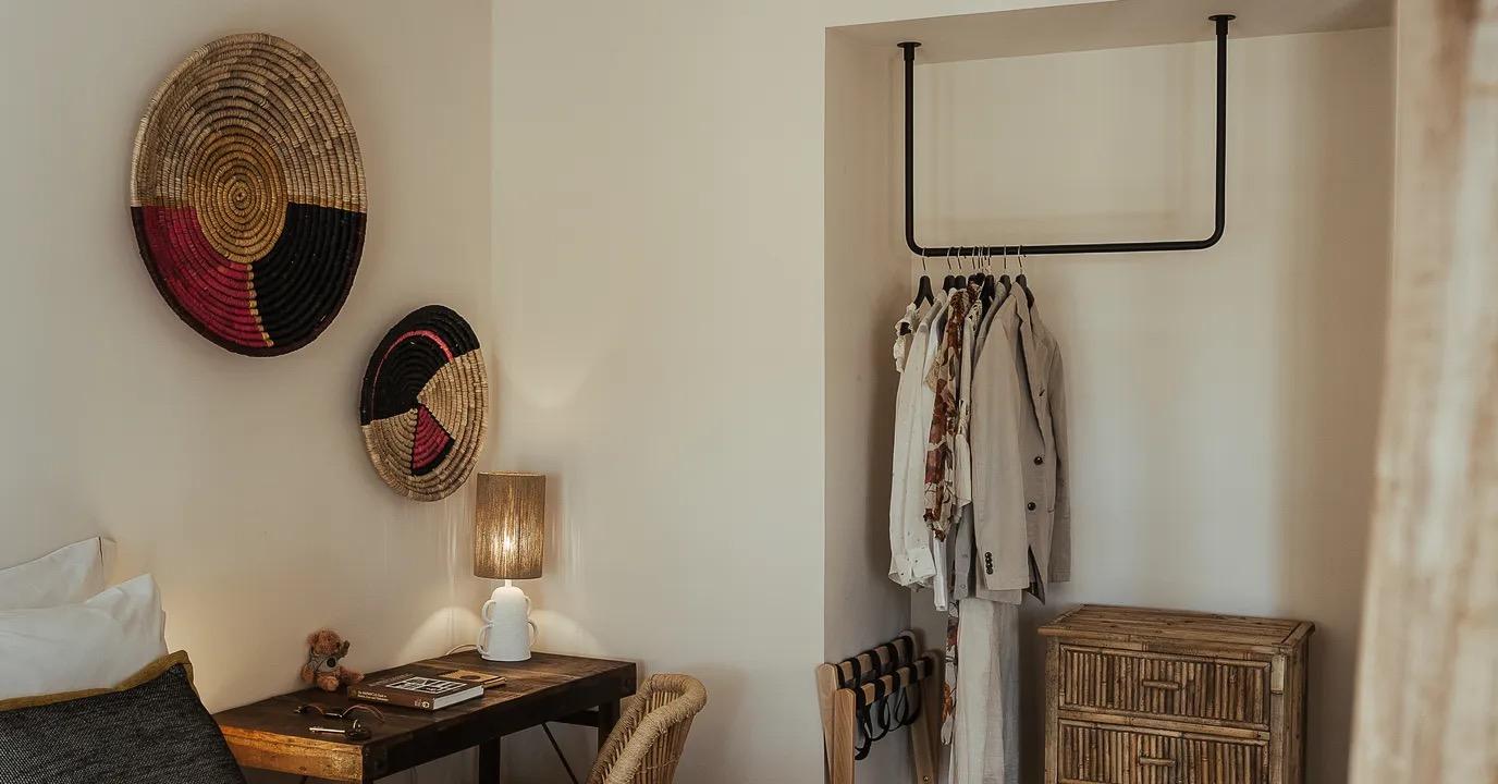 A bedroom corner with two round woven wall baskets, a wooden desk holding books and sunglasses, a cane chair, and a clothes rail hung with garments beside a rattan chest of drawers.