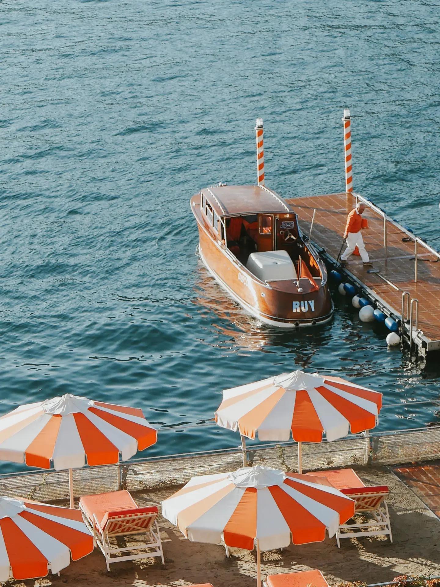 A varnished wooden motorboat named Ruy is moored at a jetty with striped mooring poles, a figure in orange walking past striped umbrellas and loungers below.