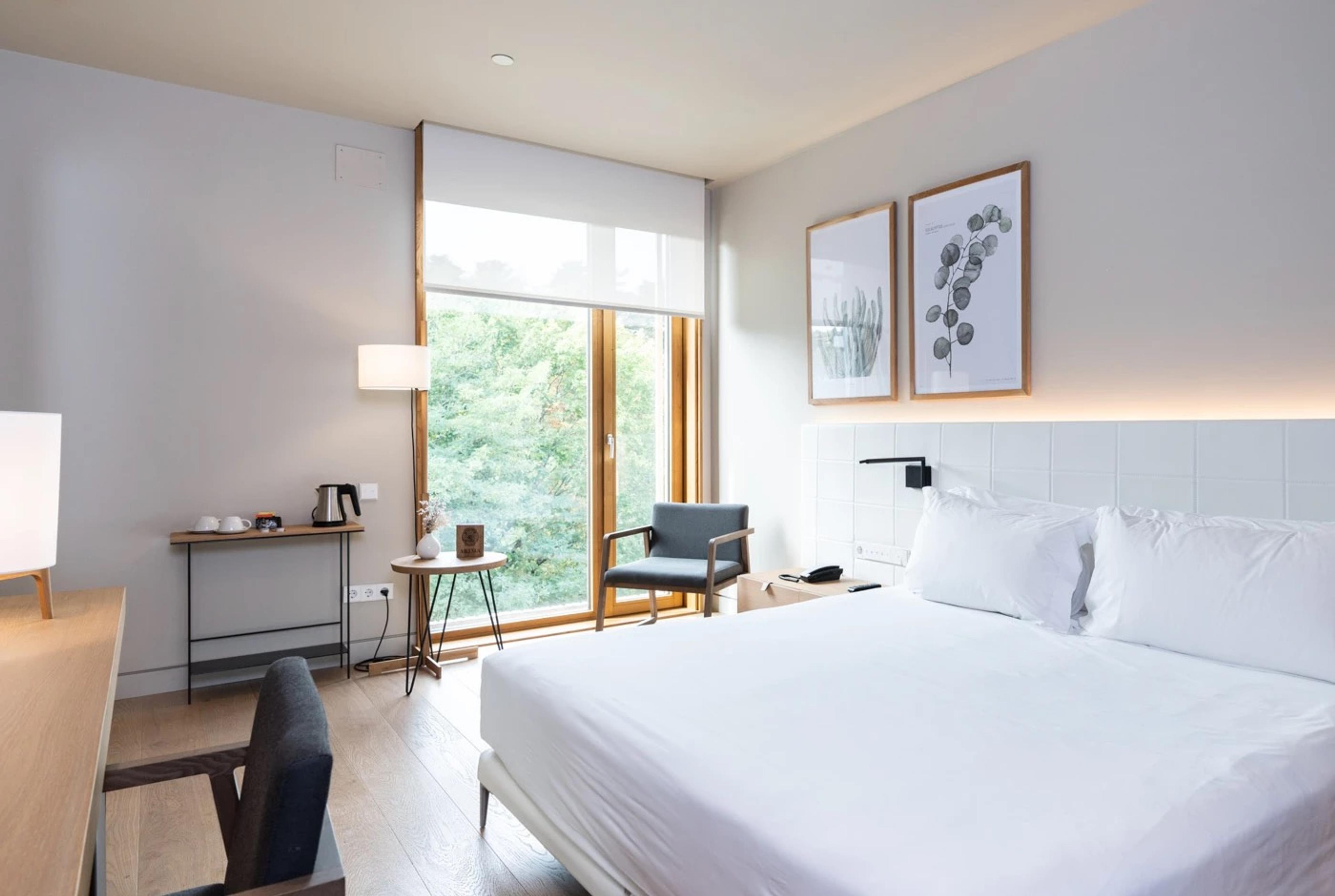 A hotel bedroom with a white bed, a desk with a kettle and a chair, and a glass door looking onto trees.