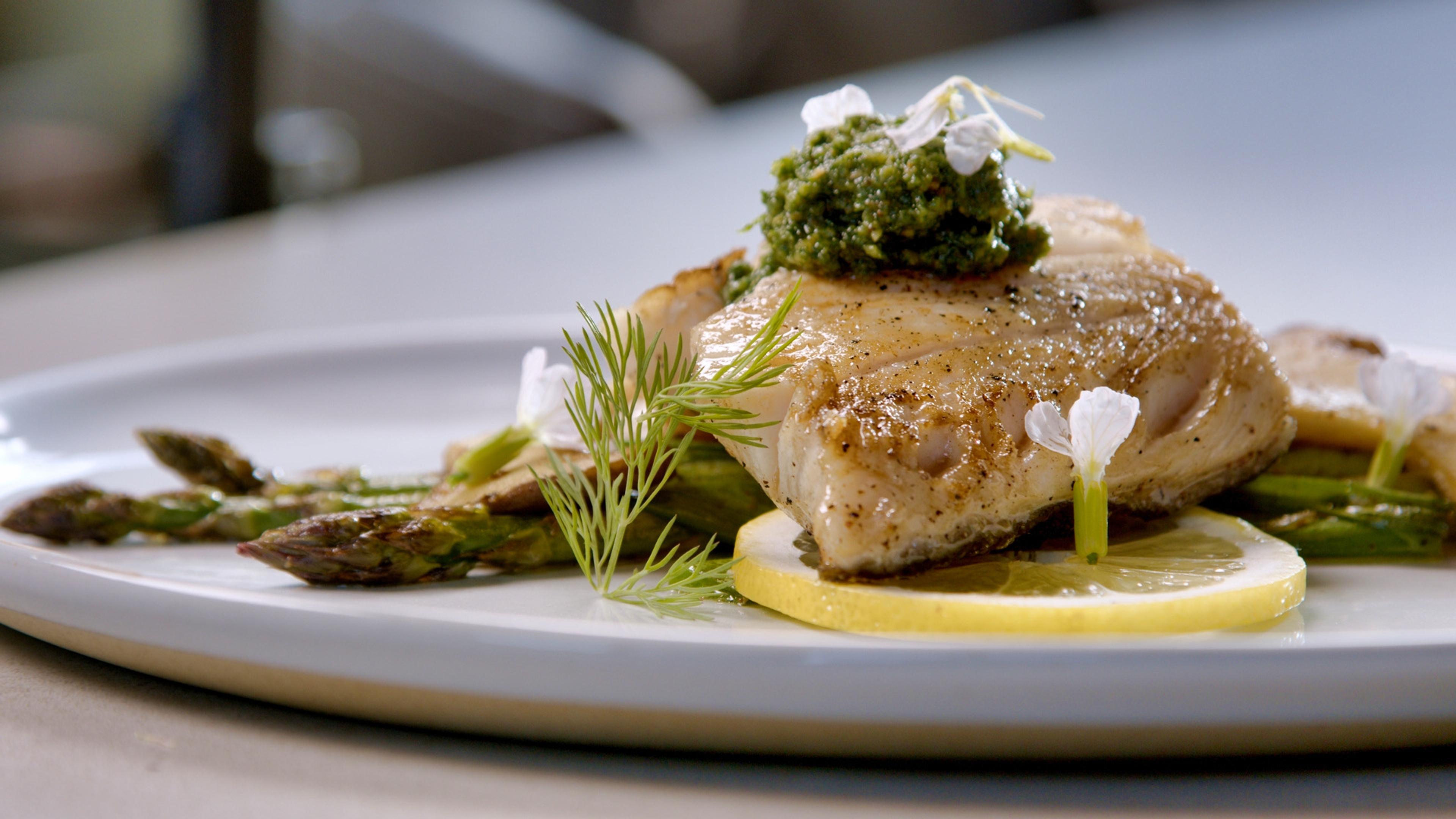 A plate of seared fish with asparagus, a lemon slice, and small white flowers on a white tablecloth.