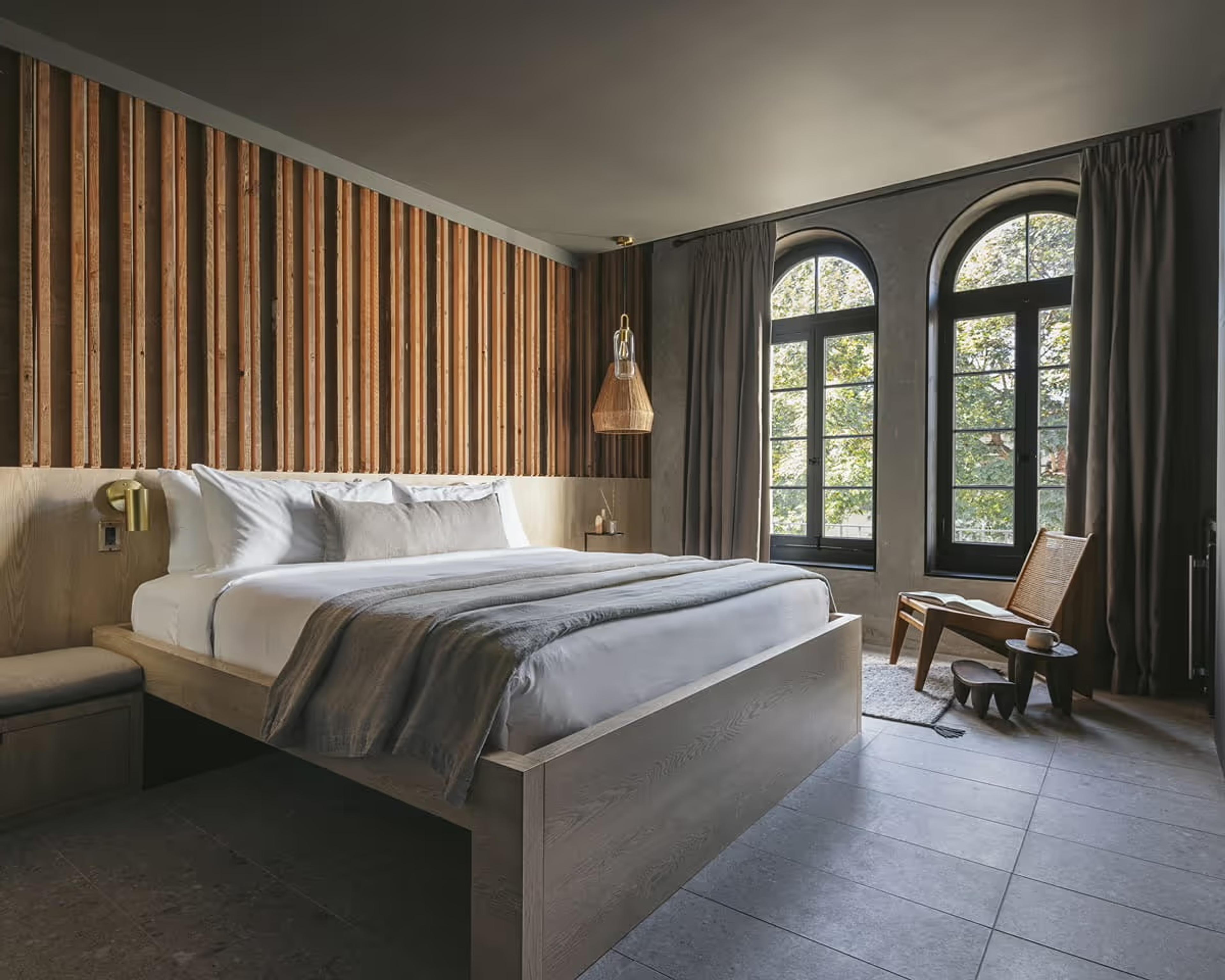 A hotel bedroom with a vertical wood slat headboard, grey and white bedding, a rattan lounge chair by two arched windows, and a woven pendant light overhead.