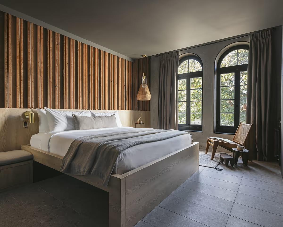 A hotel bedroom with a vertical wood slat headboard, grey and white bedding, a rattan lounge chair by two arched windows, and a woven pendant light overhead.