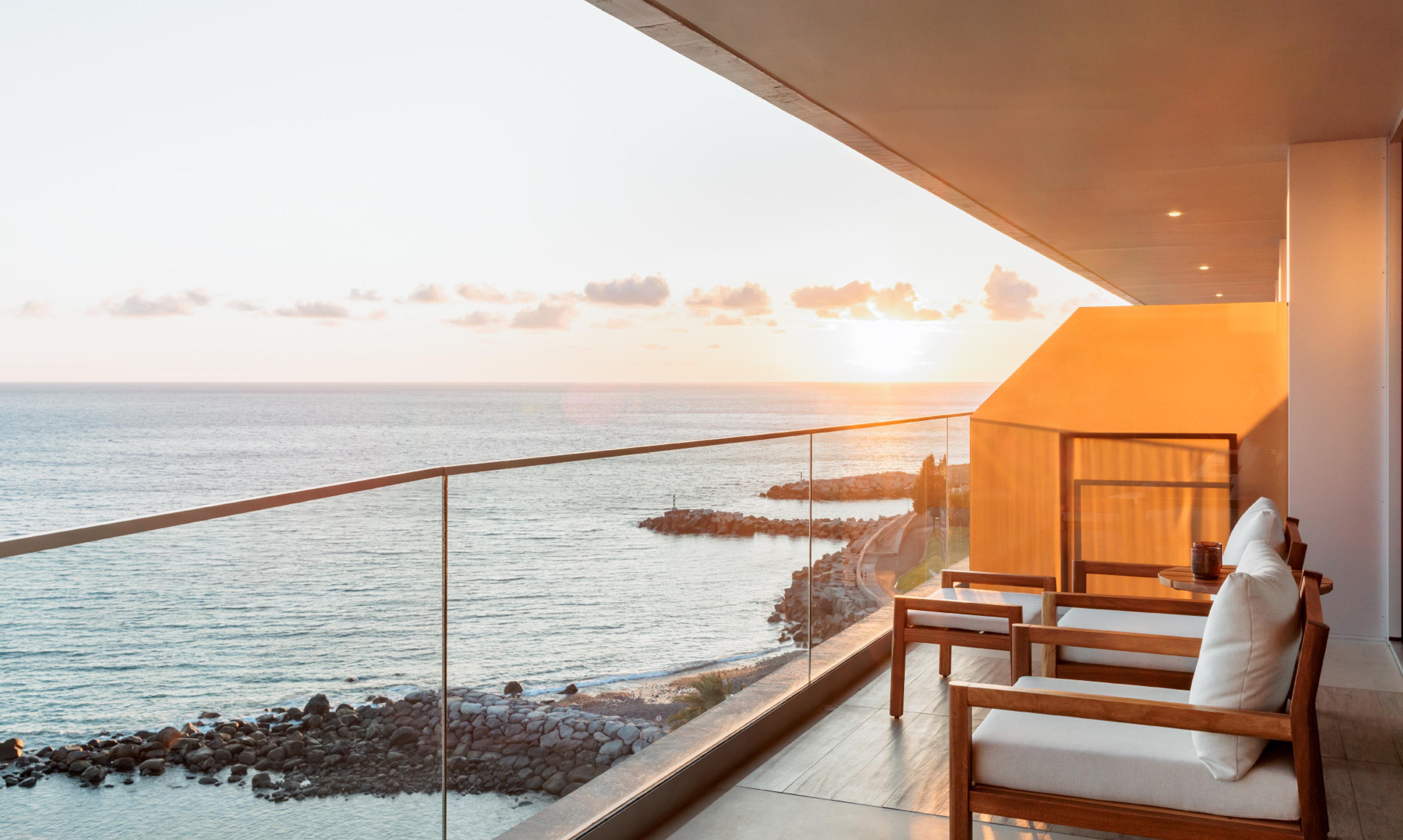 A balcony with wooden armchairs facing the sea at sunset, with a rocky coastline below.