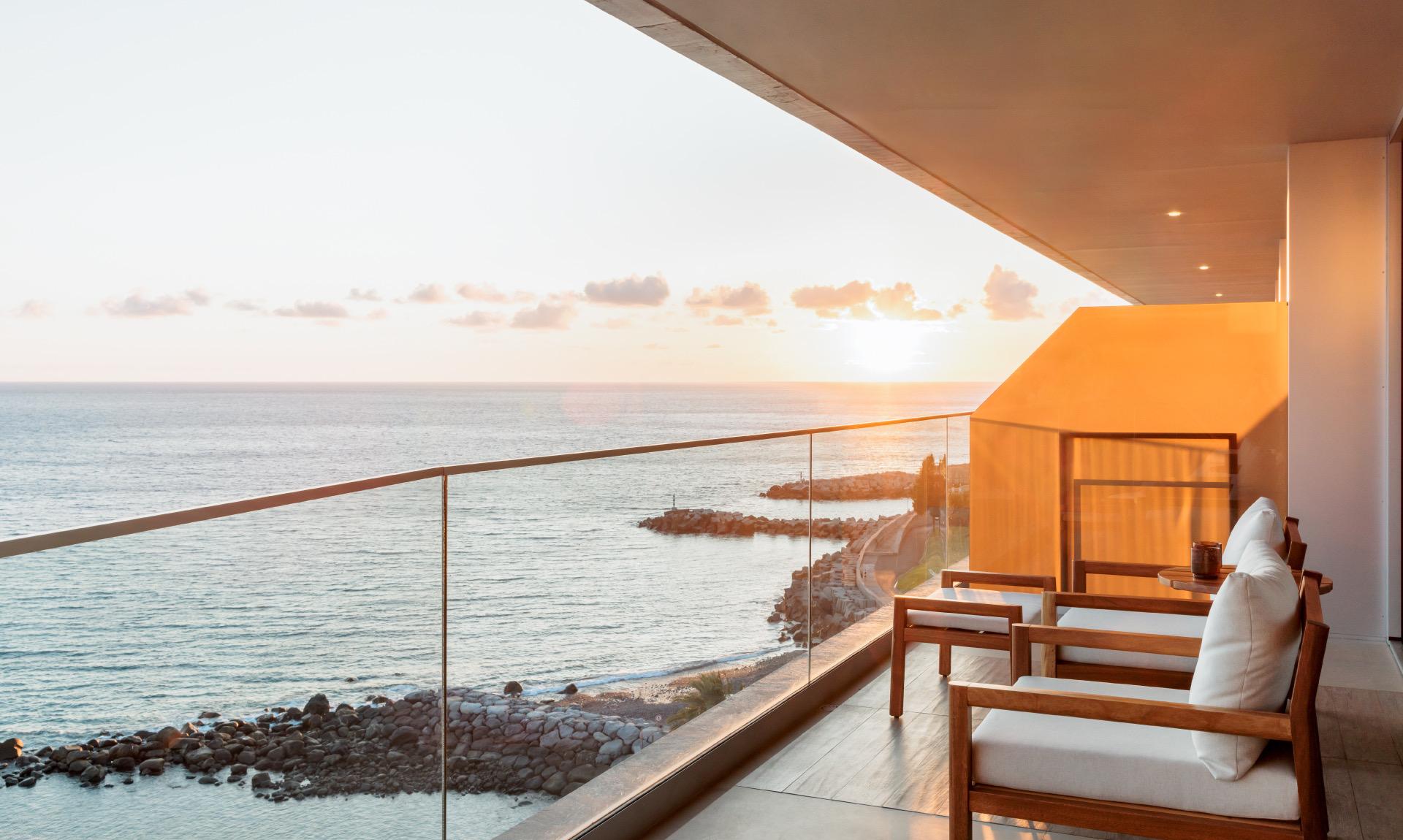 A balcony with wooden armchairs facing the sea at sunset, with a rocky coastline below.