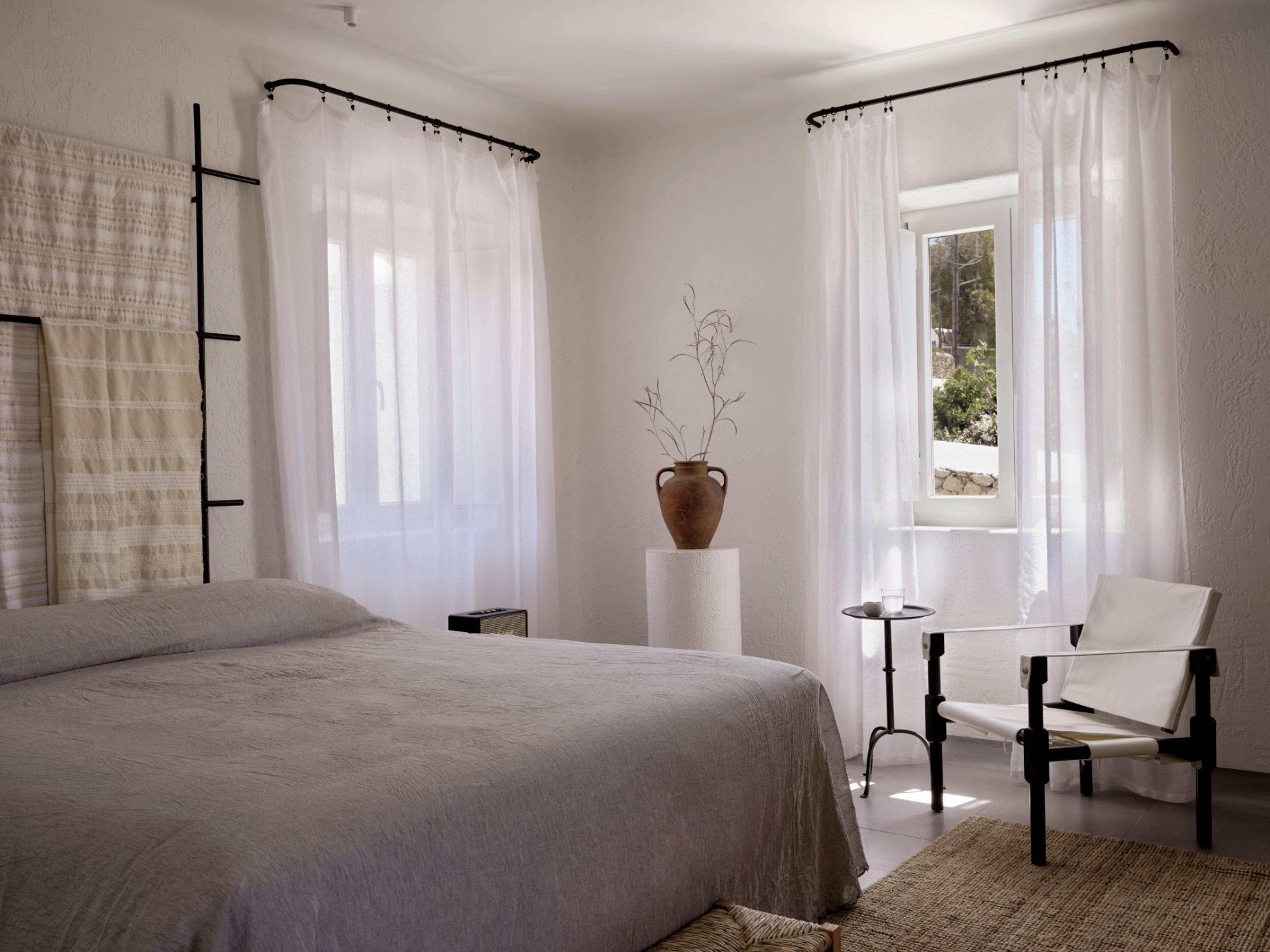 A made bed with a grey linen cover sits below a woven wall-hanging, facing two curtained windows, with a terracotta jar and a woven-seat chair in the room.