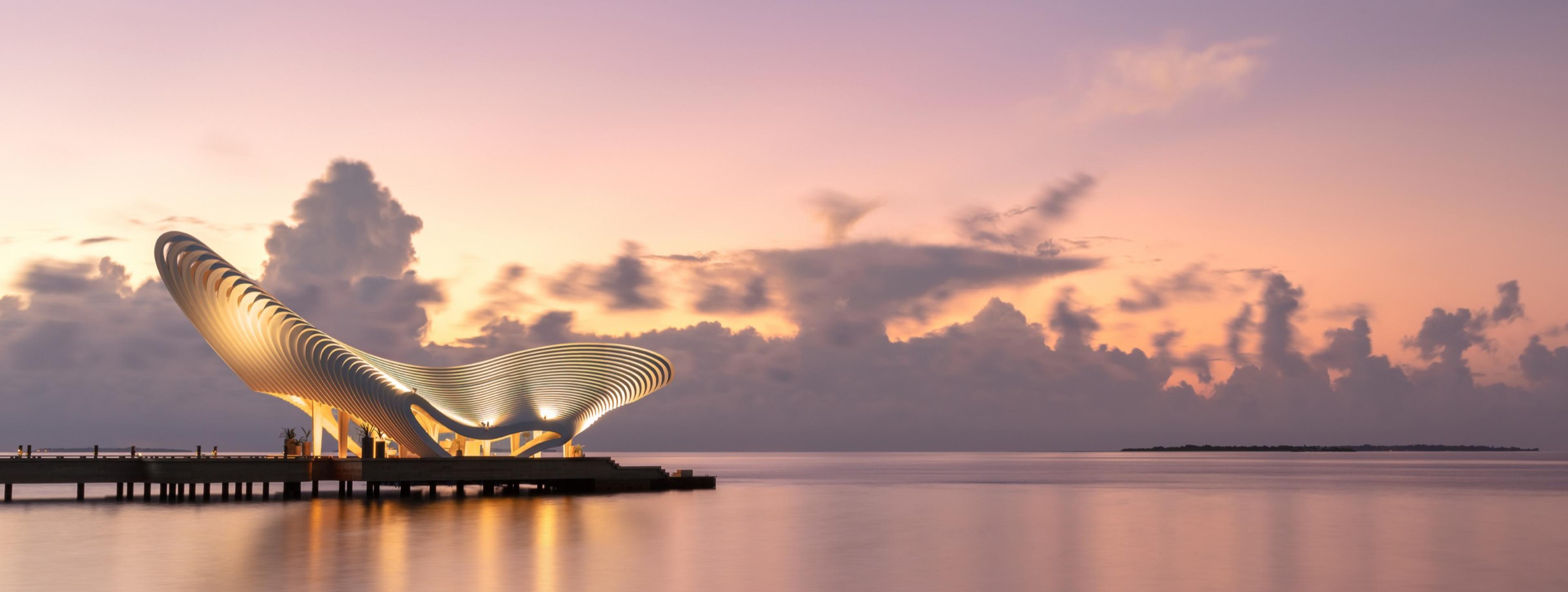 A sculpted, open-sided structure lit from within on a jetty over calm water at sunset, pink and grey clouds above.