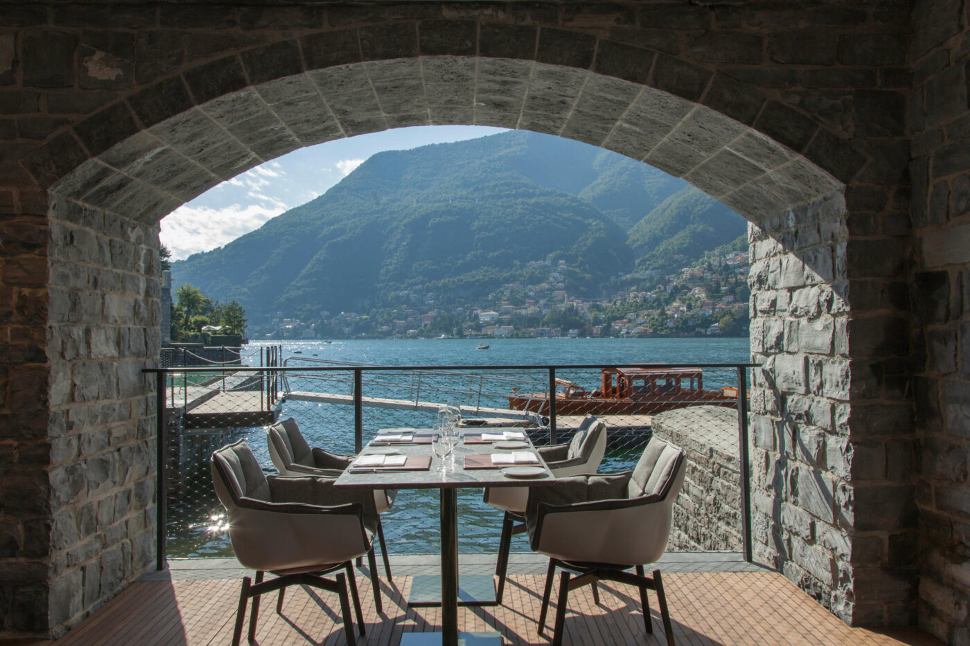 A table set for four under a stone archway on a lakeside terrace, with a wooden boat moored at a jetty in the water beyond.