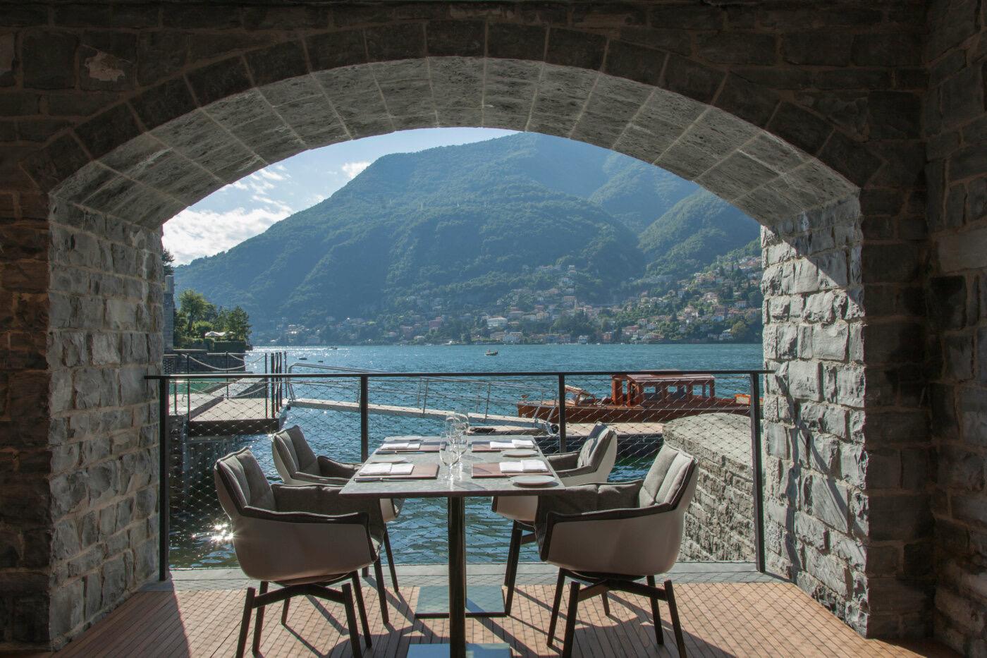 A table set for four under a stone archway on a lakeside terrace, with a wooden boat moored at a jetty in the water beyond.