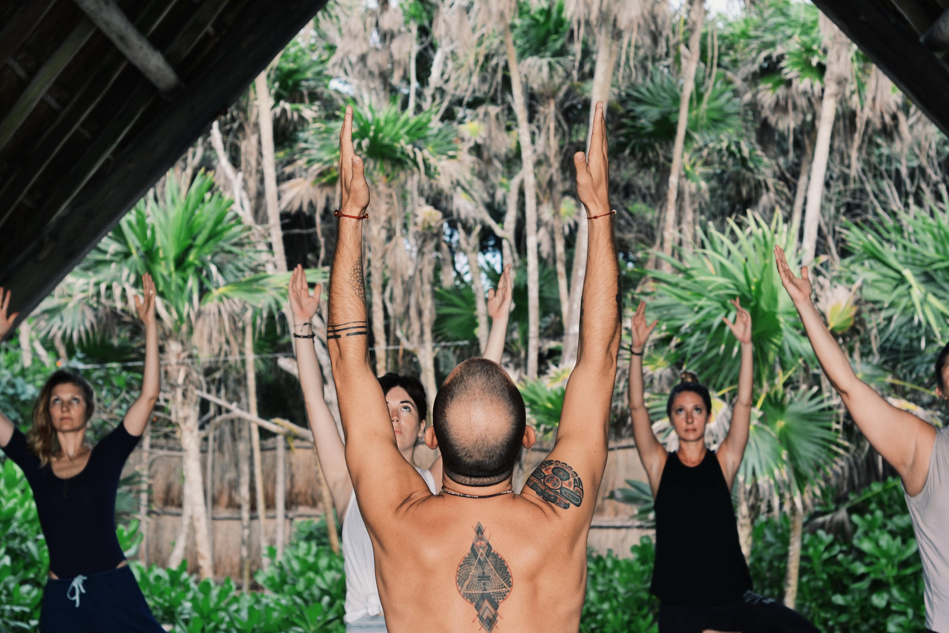 a group with arms raised in a yoga pose under a palm-thatched roof, backed by palm trees