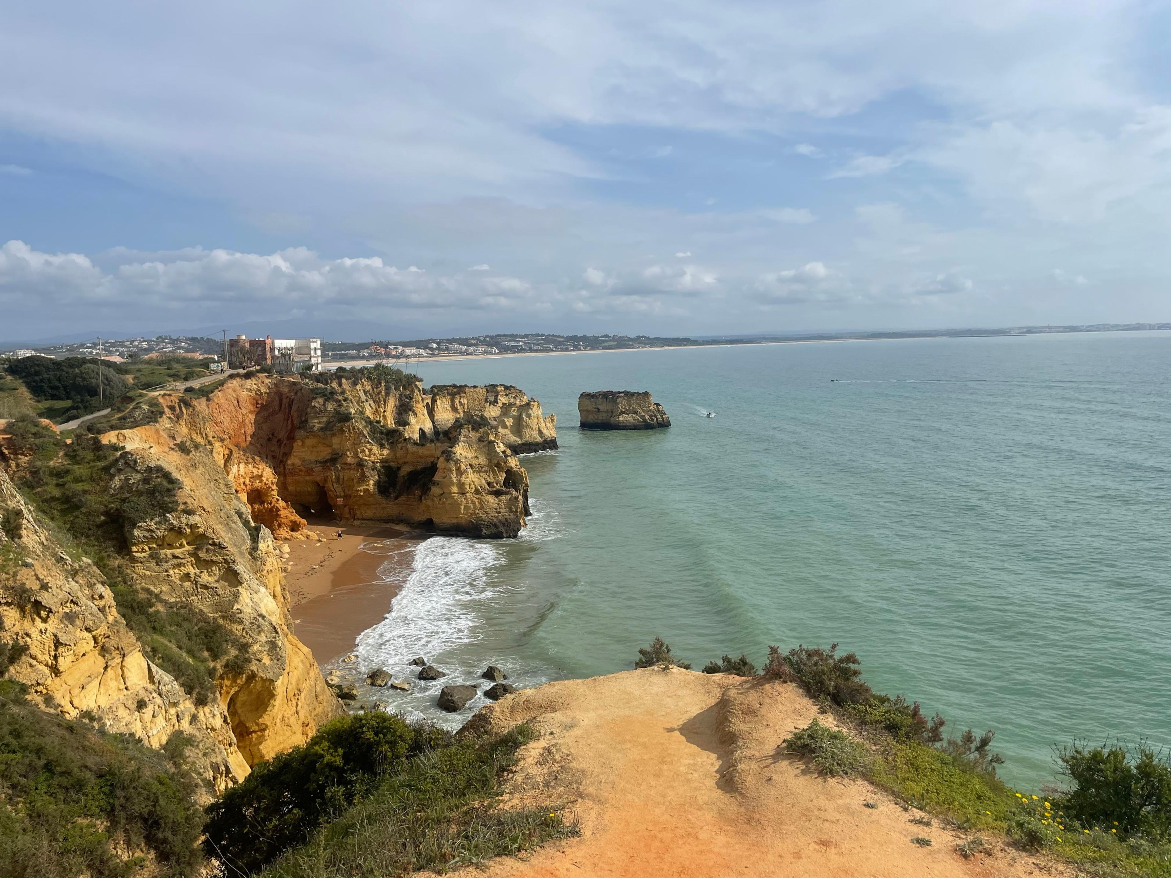 Golden sandstone cliffs and a sea stack above a small beach near Lagos, with the bay curving east behind
