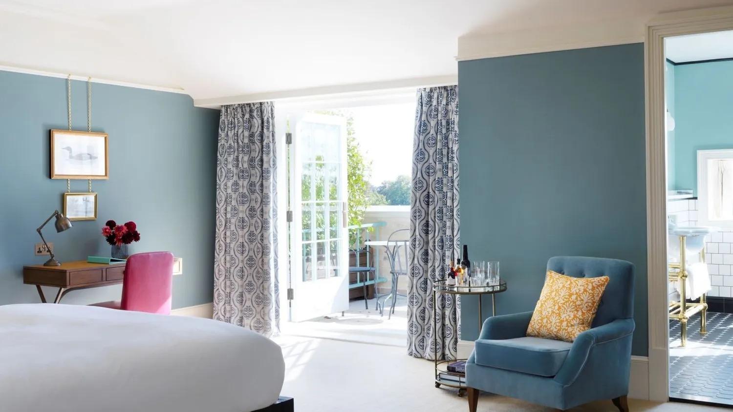 a bedroom with teal walls, a bed in the foreground, a blue armchair and a drinks trolley, and open French doors leading to a small balcony with a table and chairs