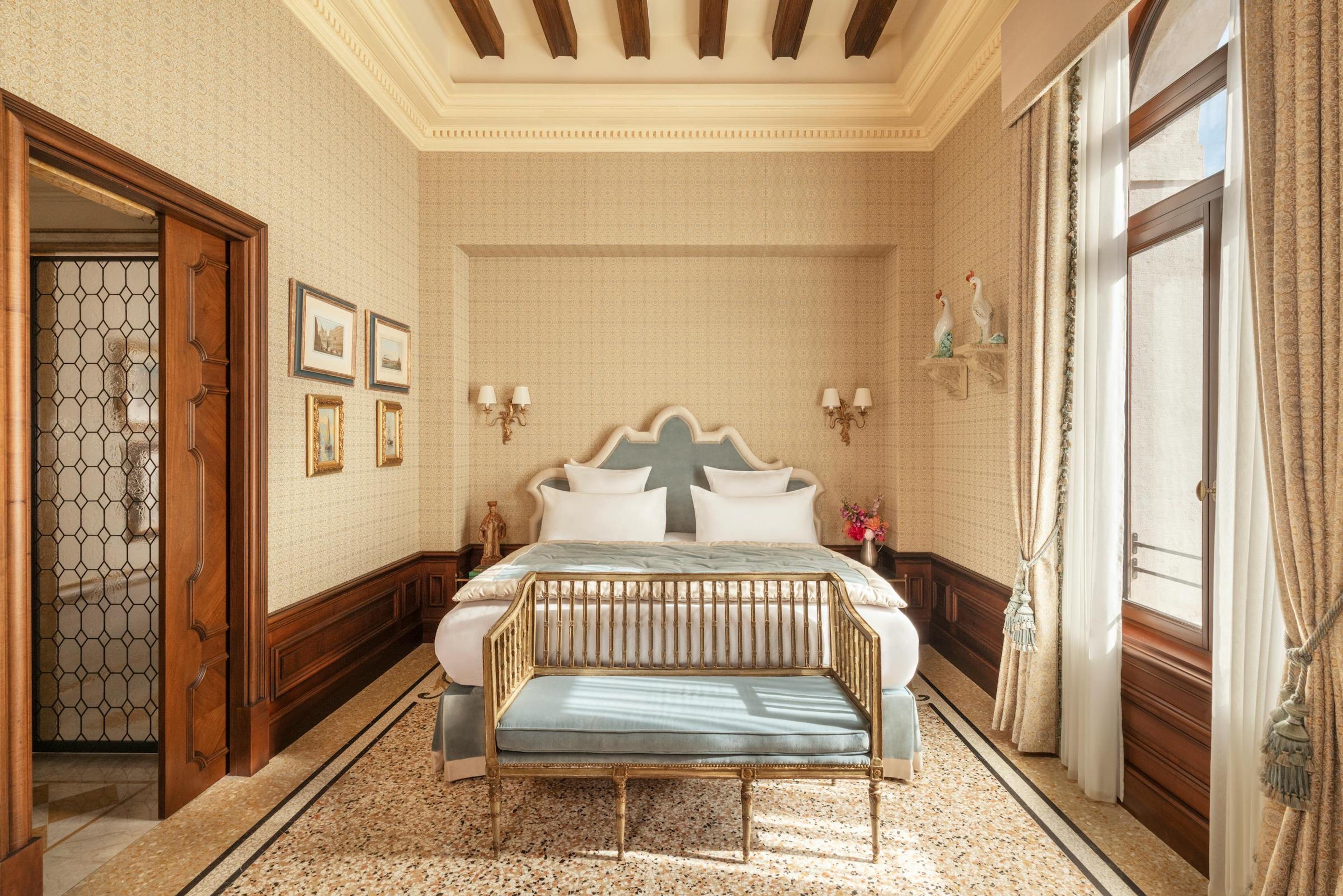 A bedroom with a pale blue upholstered headboard and bench at the foot of the bed, a terrazzo floor, framed prints on the wall, and an open door to a tiled bathroom.