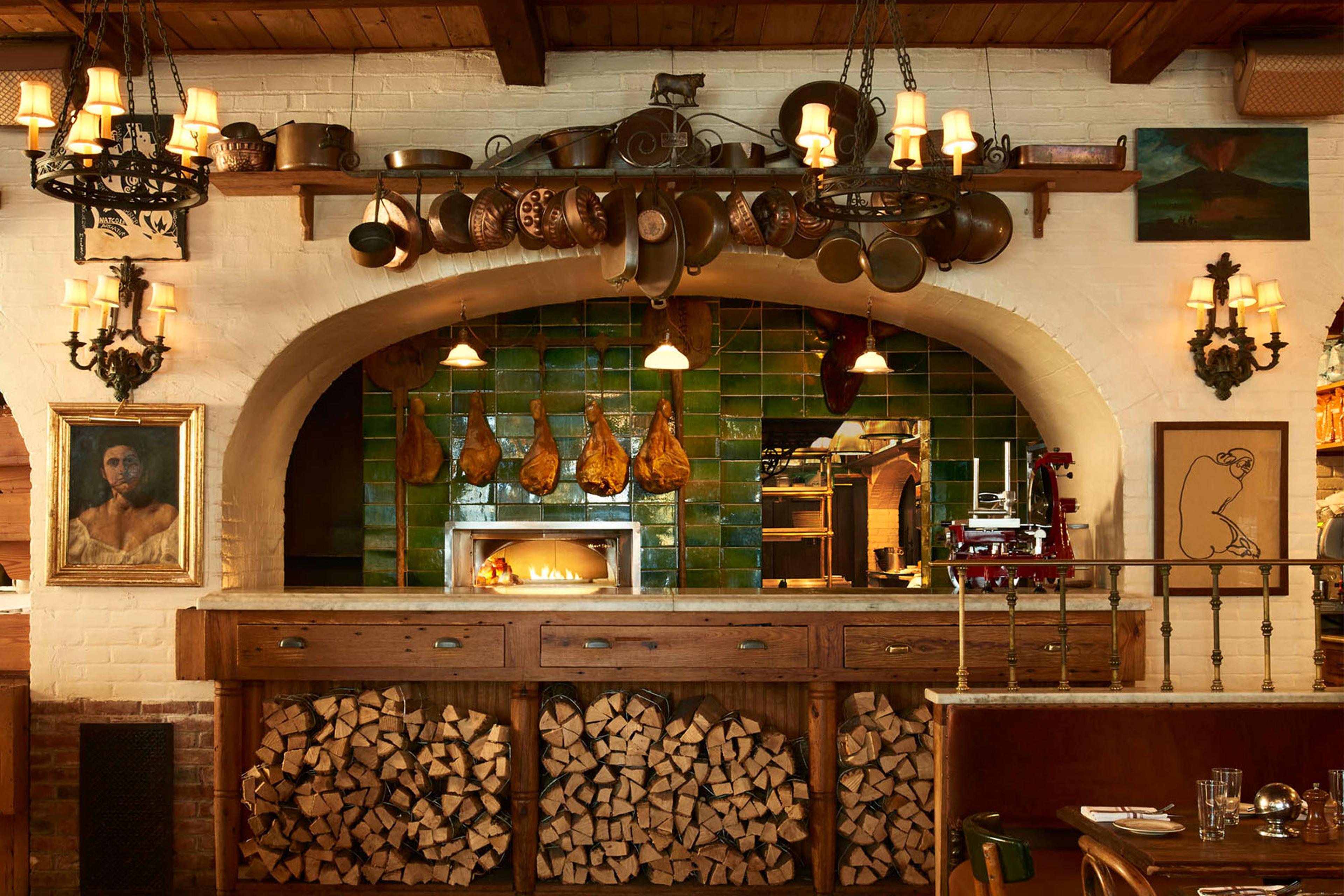 A restaurant kitchen with a wood-fired pizza oven under green tiles, hams hanging to cure, and copper pots above a shelf of wine bottles.