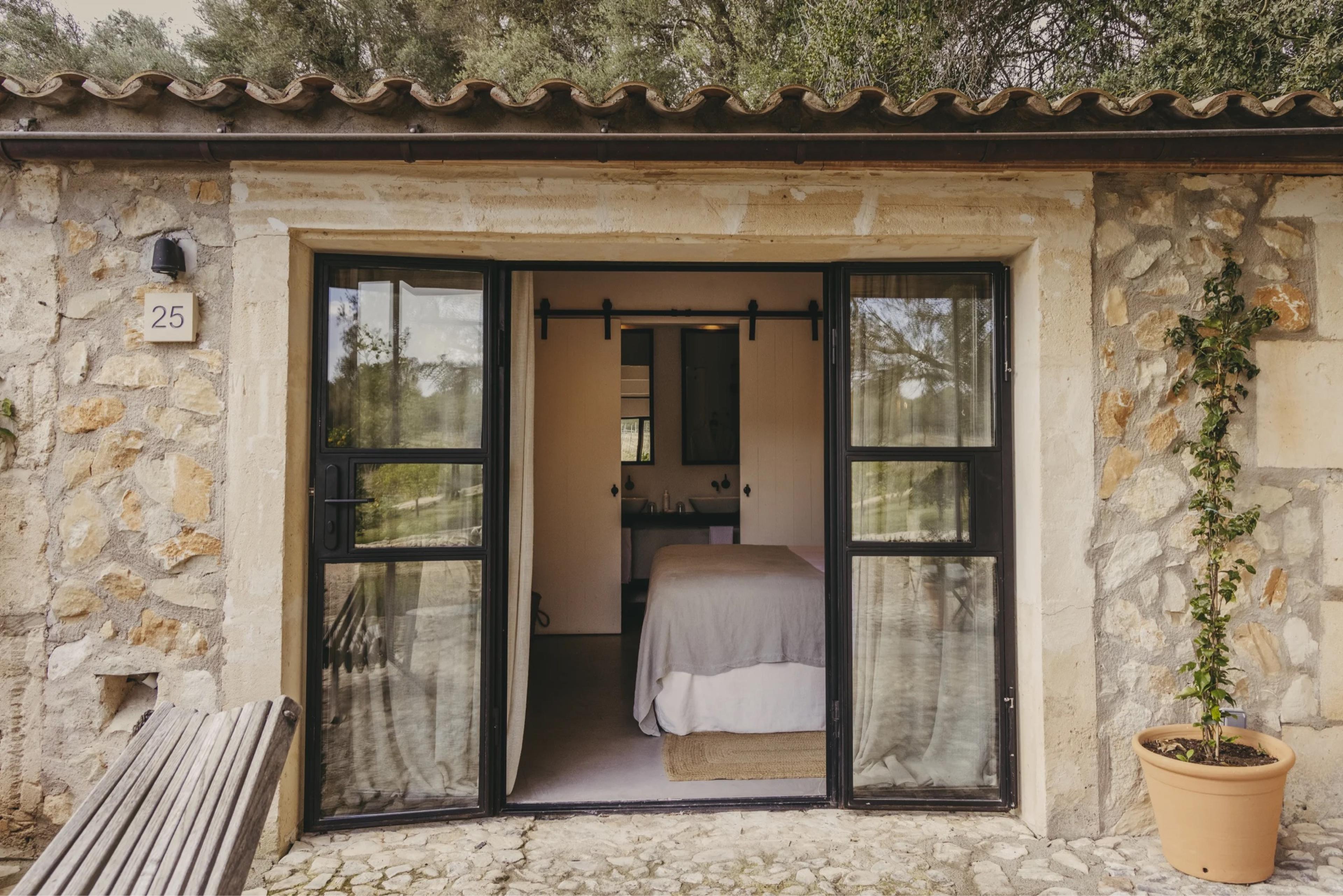 A bedroom with a made-up bed, bedside lamps, and open doors leading out to a garden.