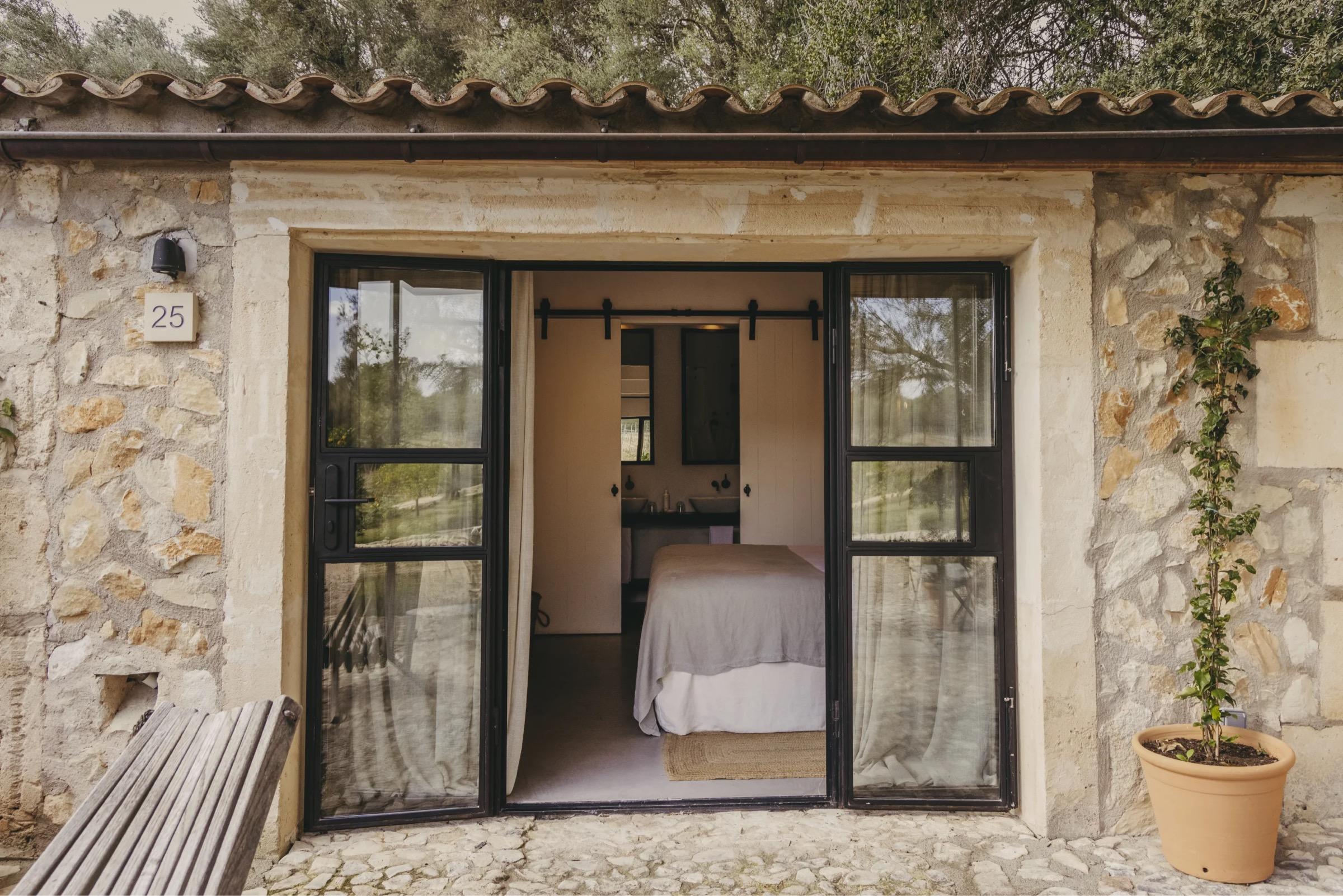 A bedroom with a made-up bed, bedside lamps, and open doors leading out to a garden.