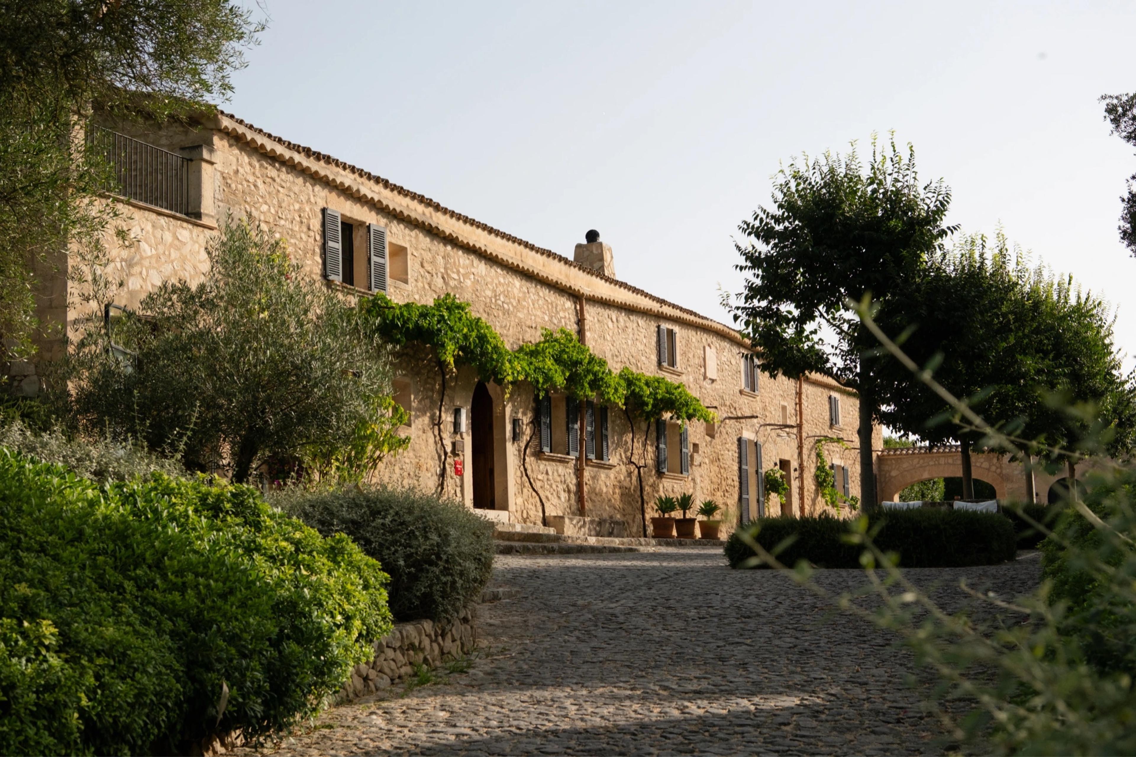 A two-storey stone farmhouse with shuttered windows and a vine growing along its facade, seen from a cobbled courtyard.