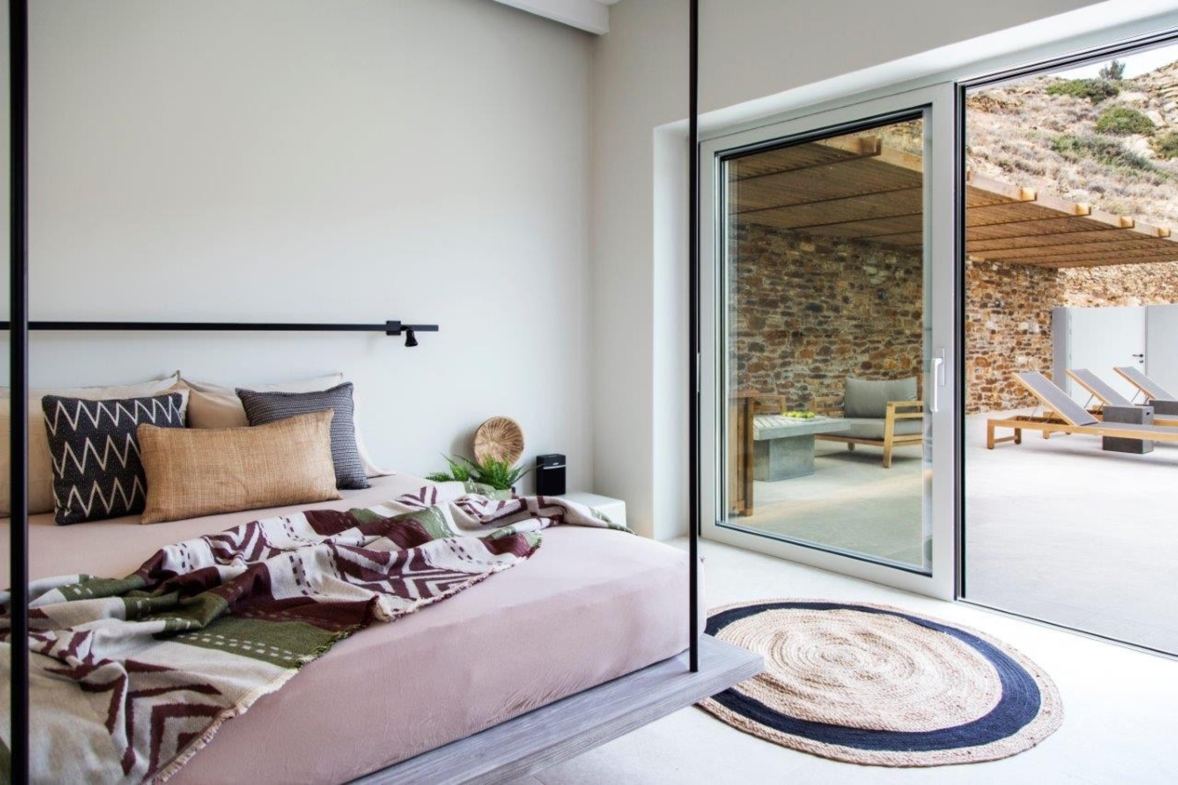 A bedroom with a pink bed, a patterned throw and a black frame, beside sliding glass doors onto a stone terrace with loungers.