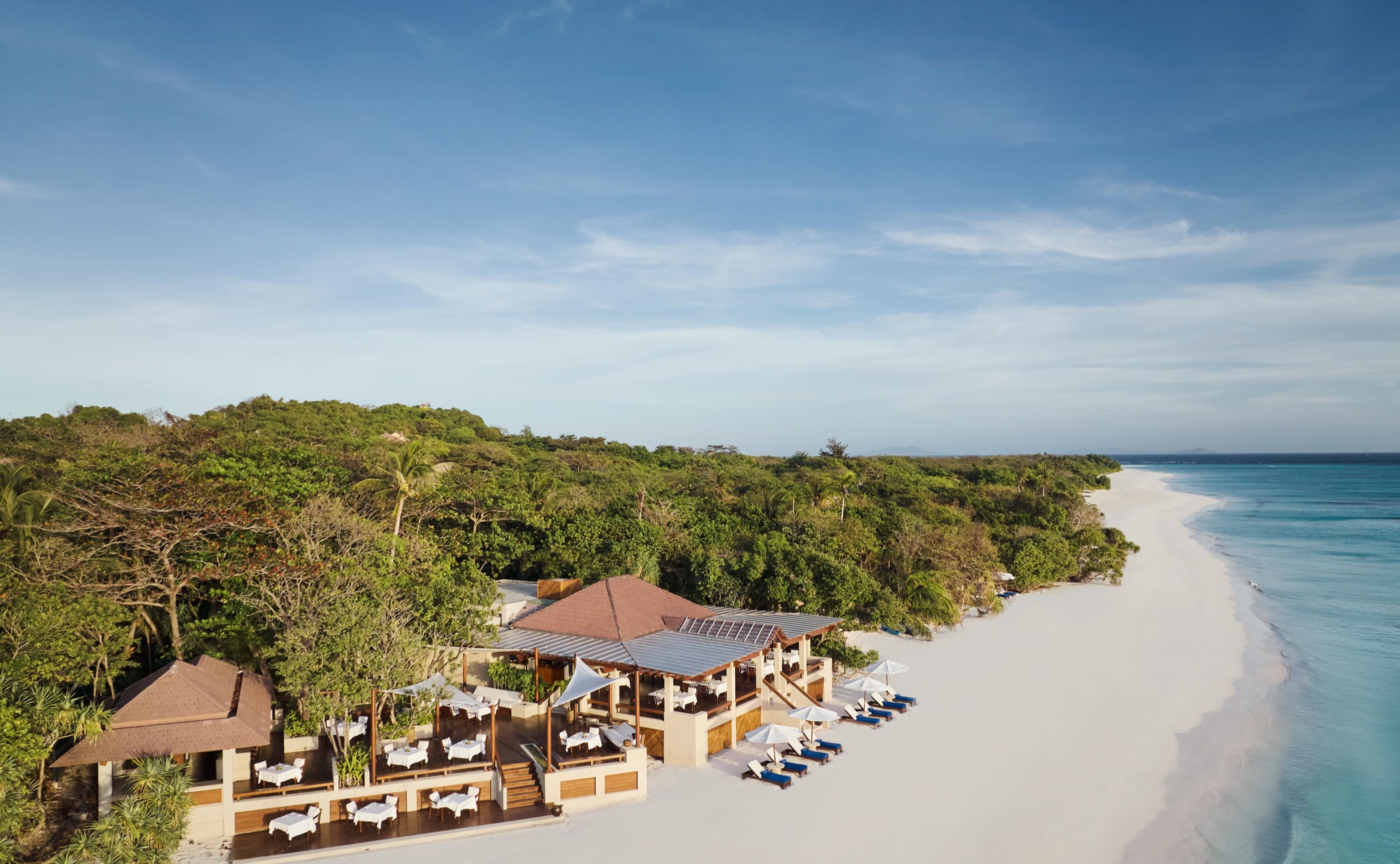 An aerial view of a jungle covered island fringed by white sand and a reef shading from turquoise to deep blue, with the resort's pavilions set back among the trees and a boat moored offshore.