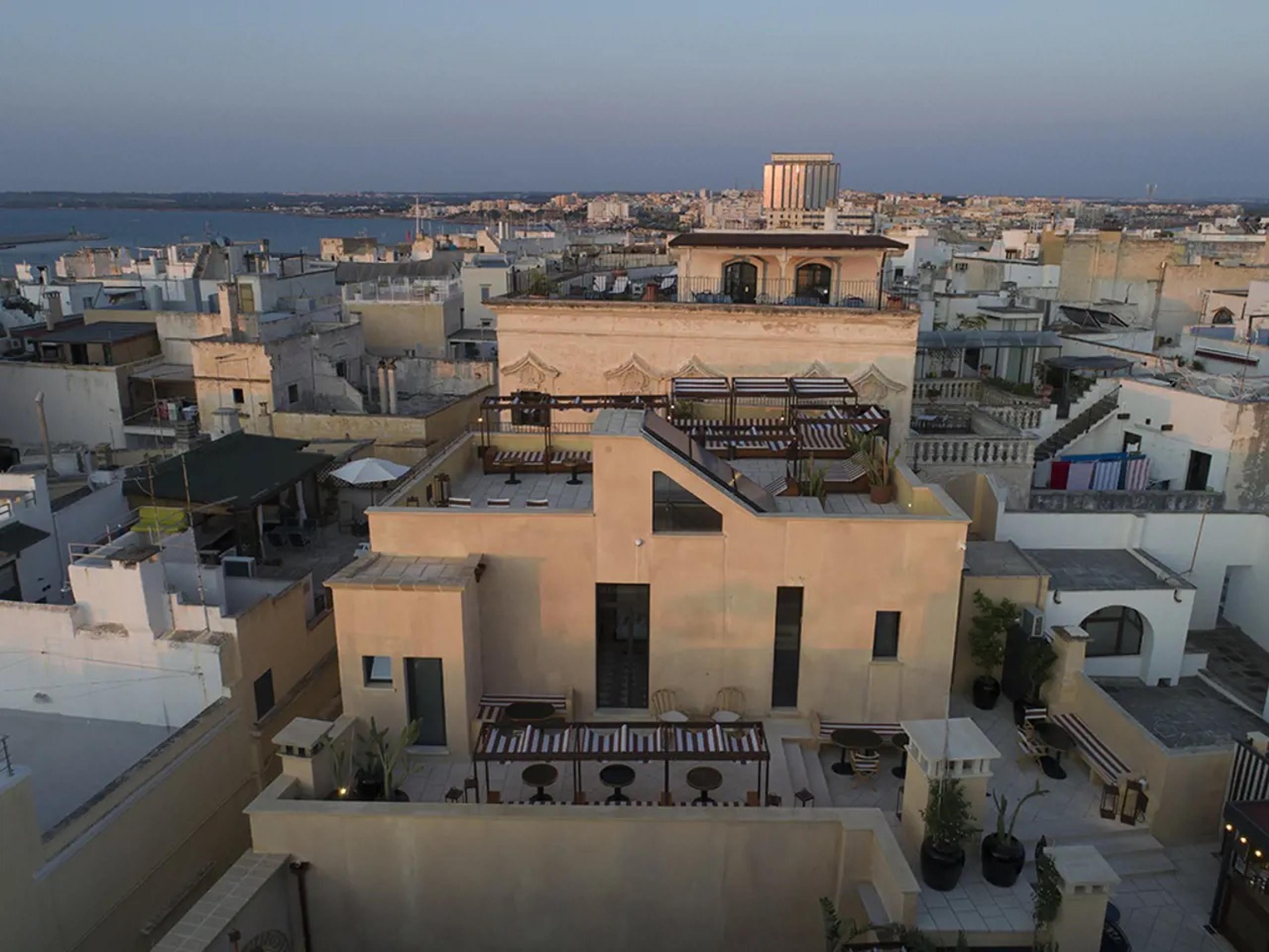 An elevated view of the hotel's pale ochre building and rooftop terrace, with striped loungers, potted cacti and seating areas, surrounded by the town's rooftops and the sea beyond at dusk.
