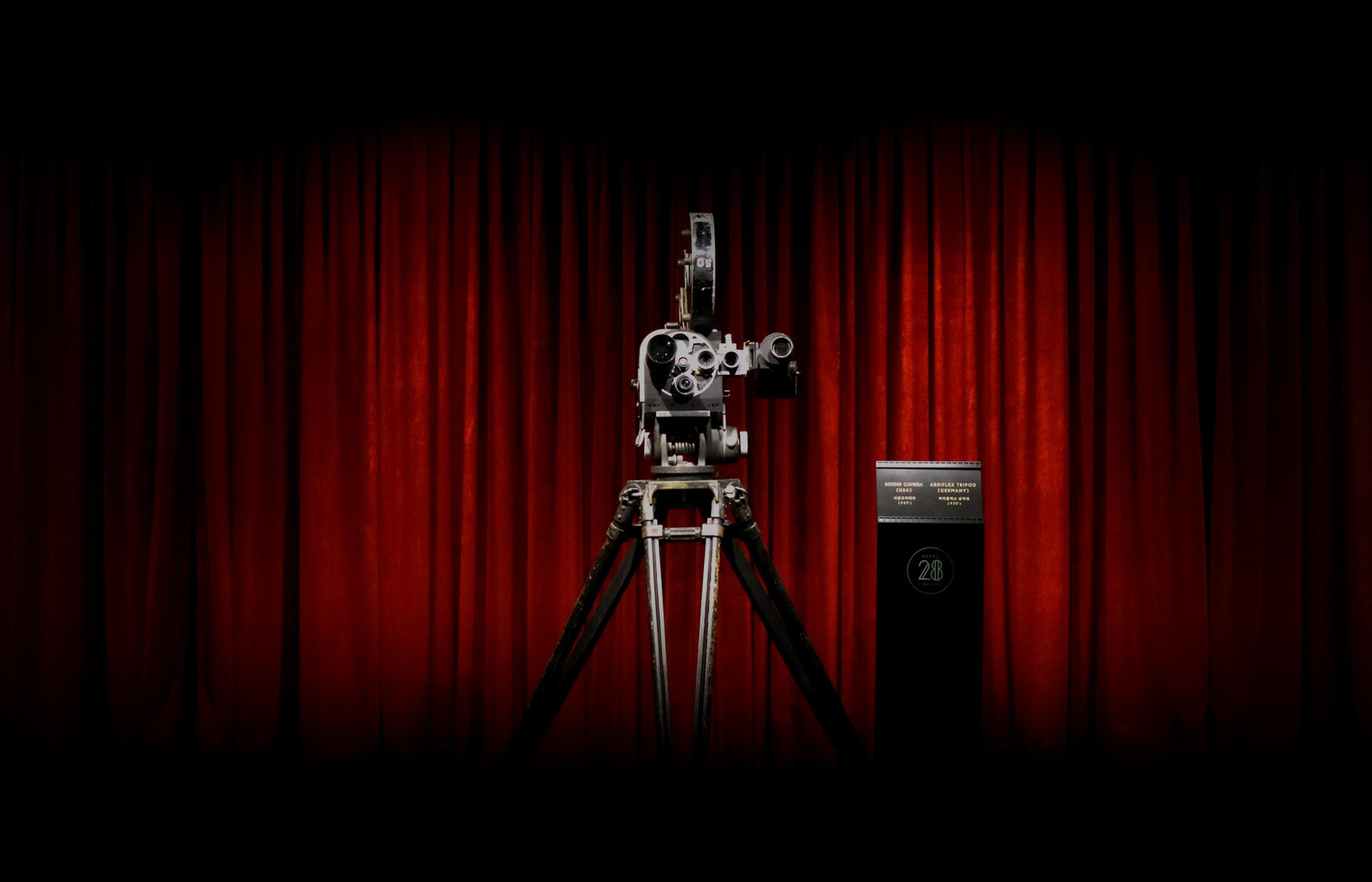 An old film camera on a tripod lit against a red curtain, with a small illuminated plaque reading 28 beside it.