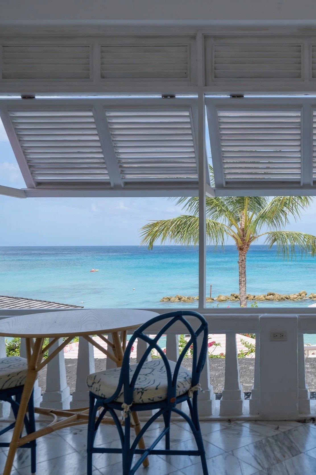 a table and chair on a shaded terrace looking out past a palm tree to the open sea