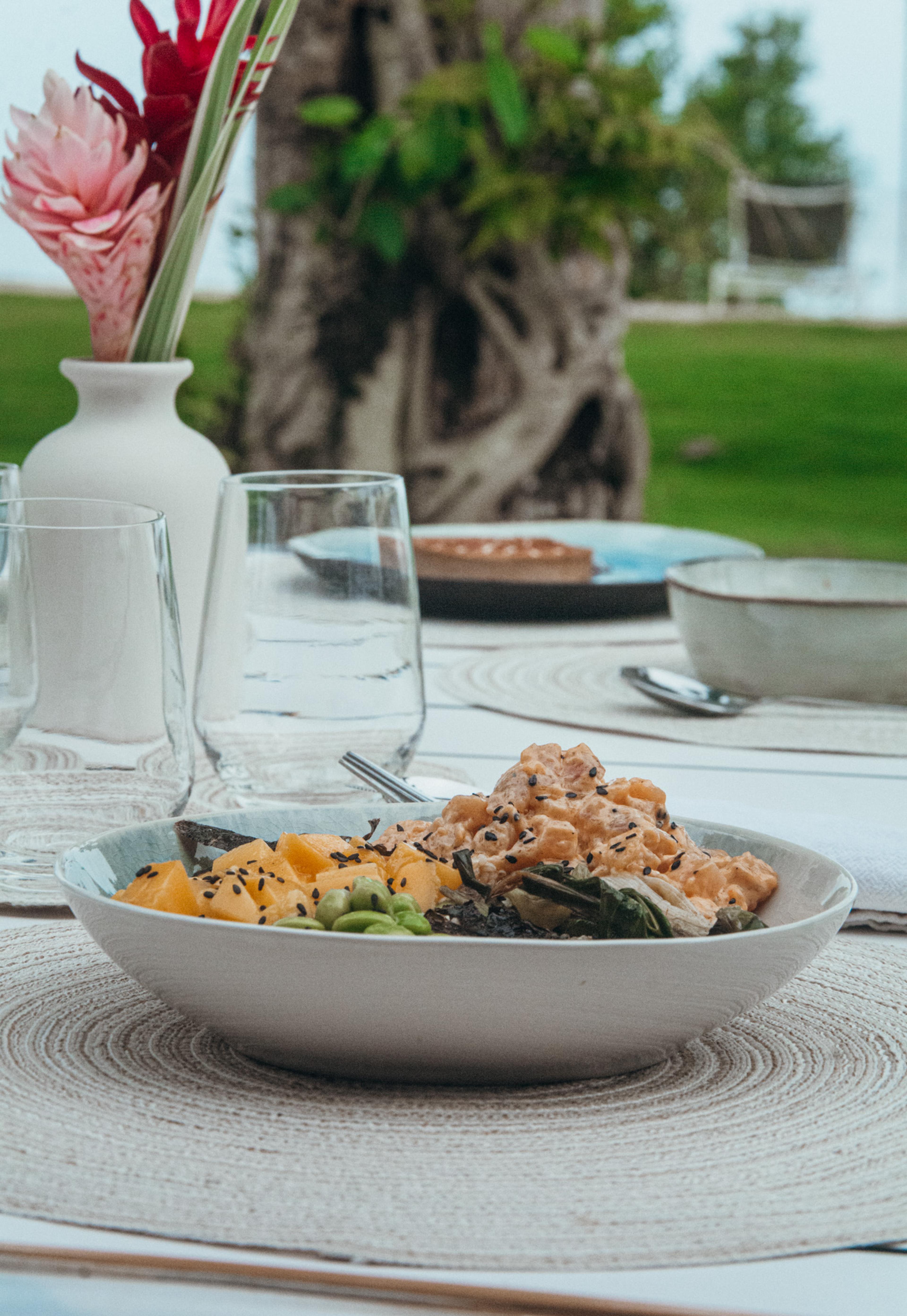 A bowl of poke with mango, edamame and seaweed sits on a table laid with wine glasses and a vase of ginger flowers.