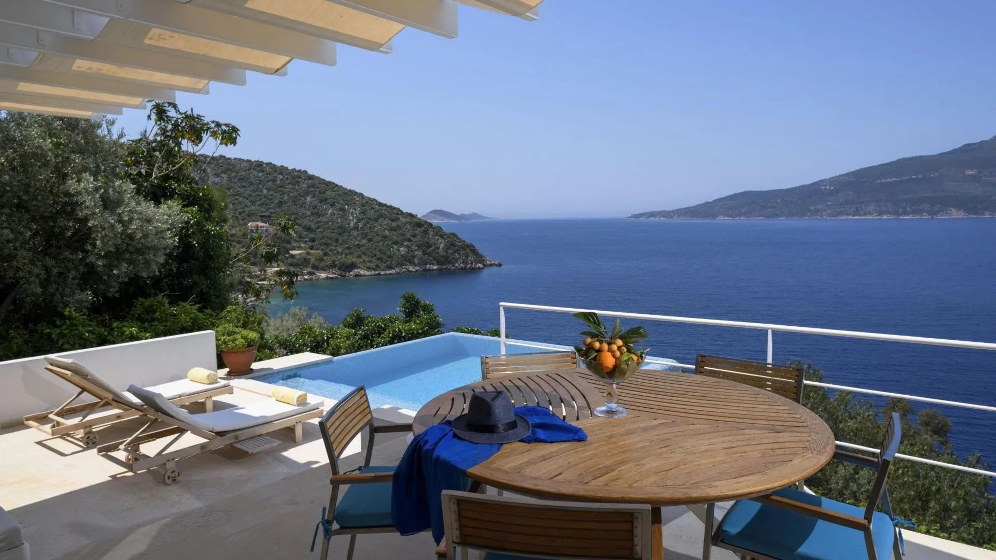 A round teak table on a terrace with a blue hat and a glass bowl of oranges on it, a pool and sun loungers at the edge, and the sea and hills beyond.