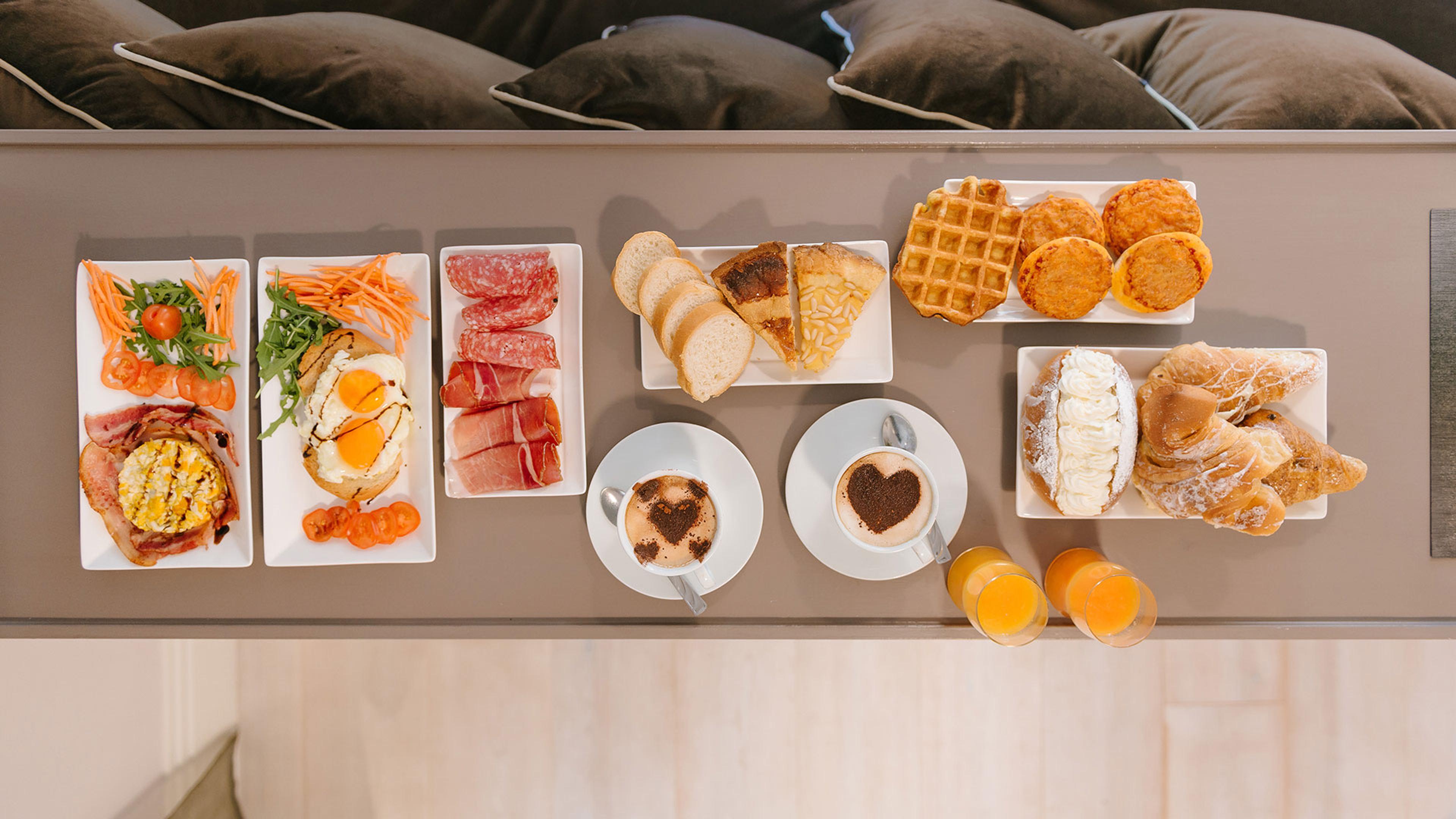 An overhead view of a breakfast spread laid on a table, with eggs, cured meats, salad, bread, a waffle, pastries, two cappuccinos and two glasses of orange juice.