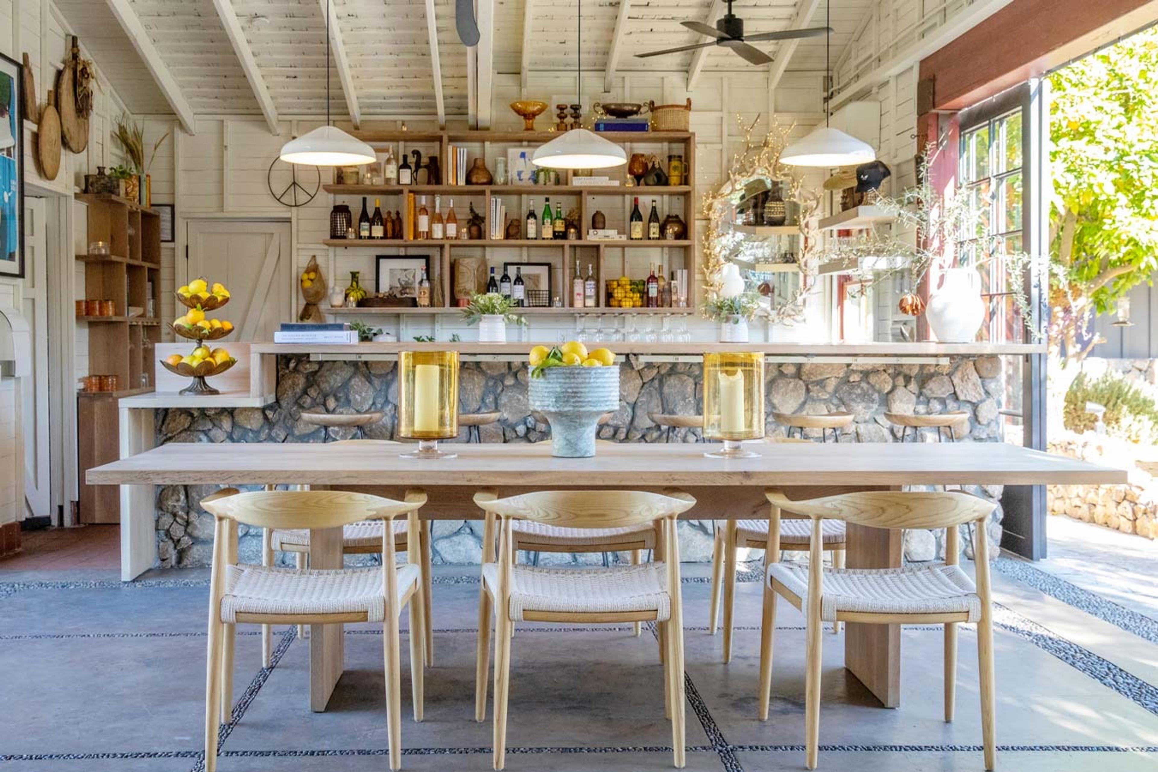 A long pale wooden dining table with pale wooden chairs in a barn-style kitchen, under white pendant lights, with a stone-faced bar and shelves of bottles behind it.