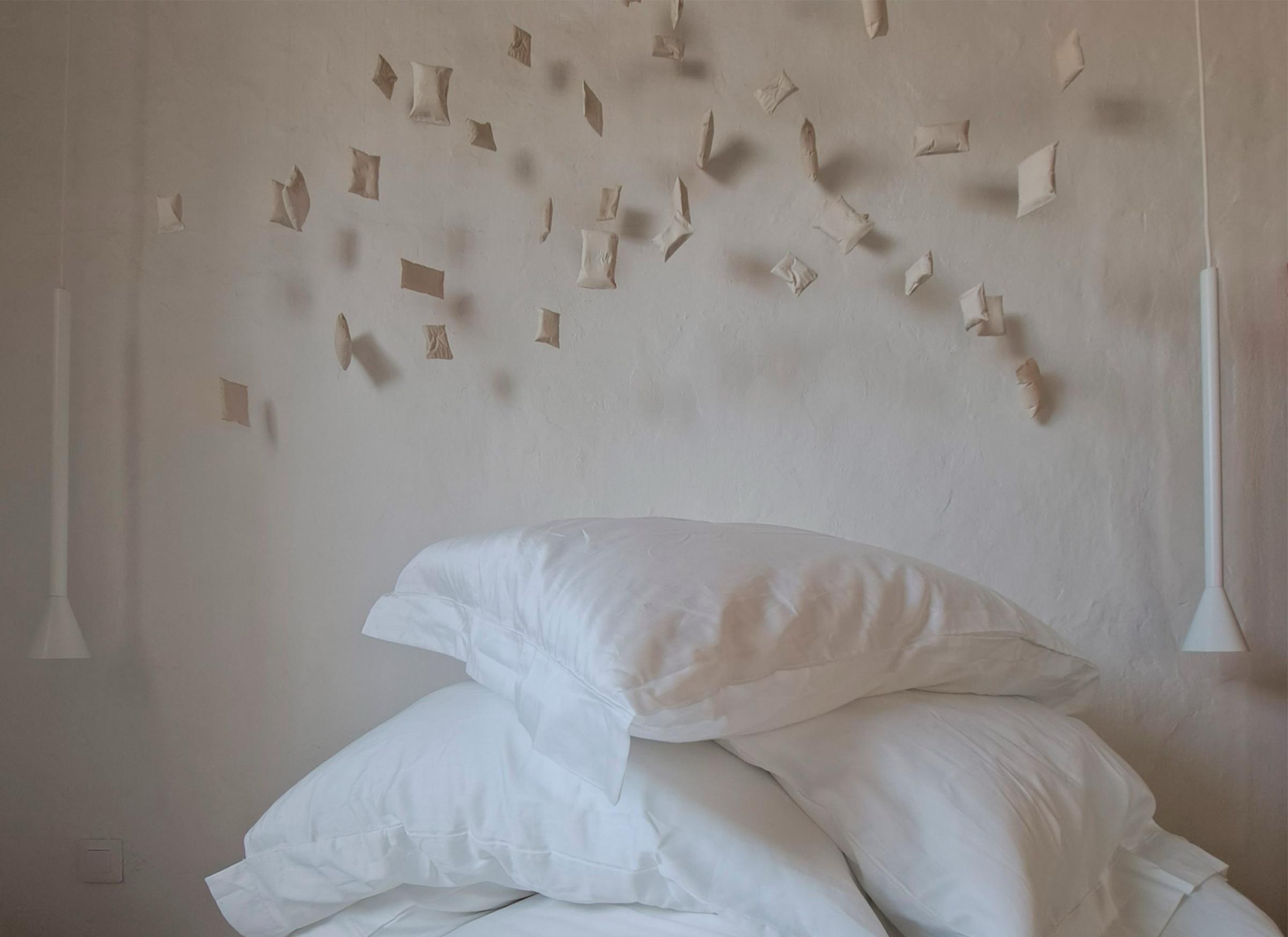 A stack of white pillows on a bed beneath a plastered wall hung with small raised paper or clay shapes, flanked by two cone-shaped pendant lights.