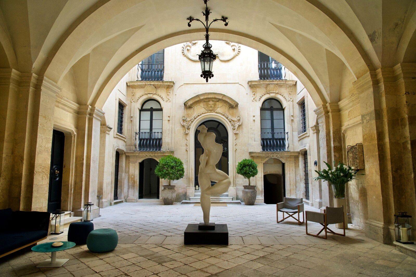 An arched stone loggia opening onto a palazzo courtyard, with a nude female statue on a plinth, potted plants, armchairs, and a hanging lantern.