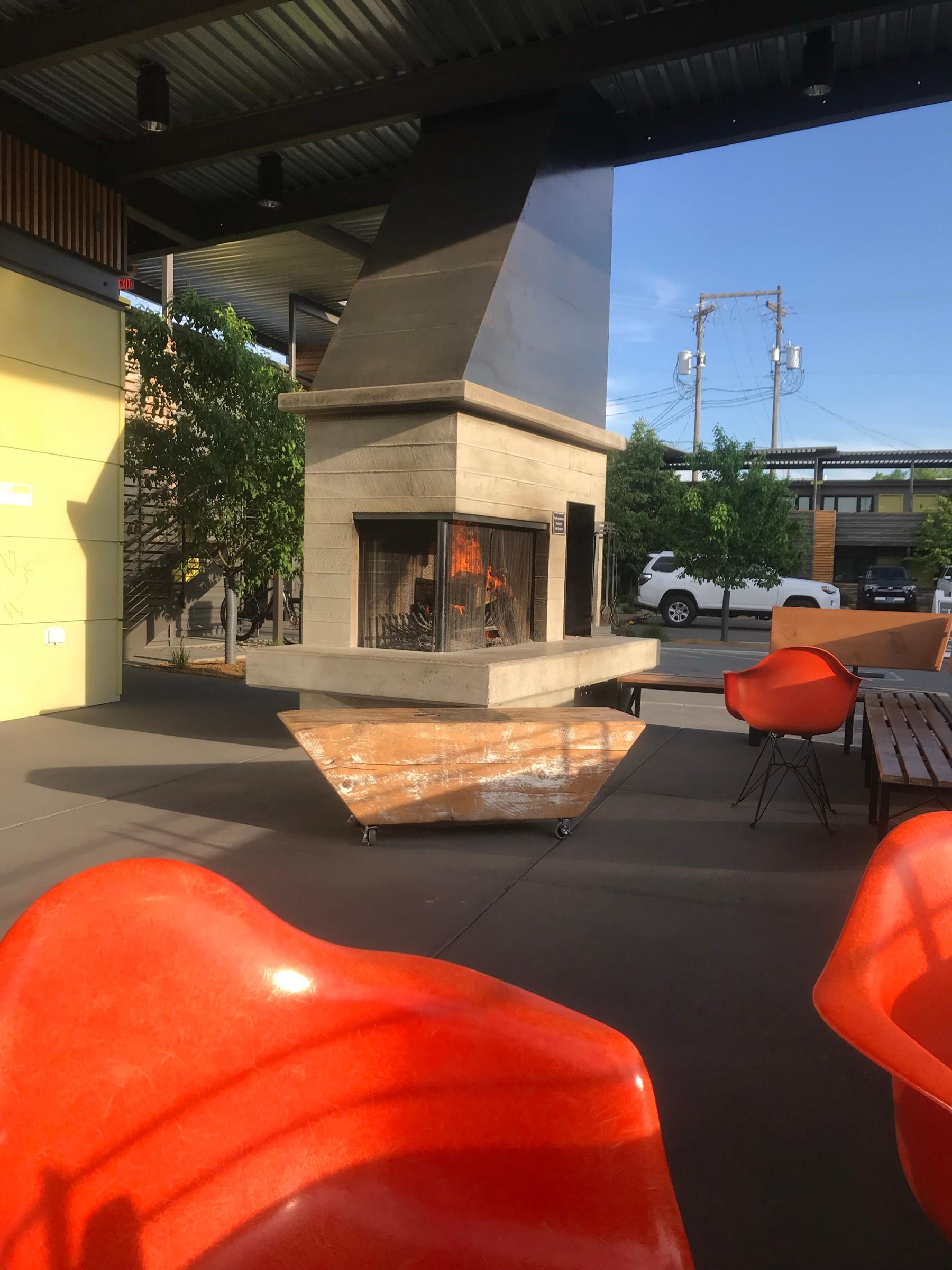 An outdoor fireplace of pale concrete under a steel hood, with orange chairs drawn up on the terrace