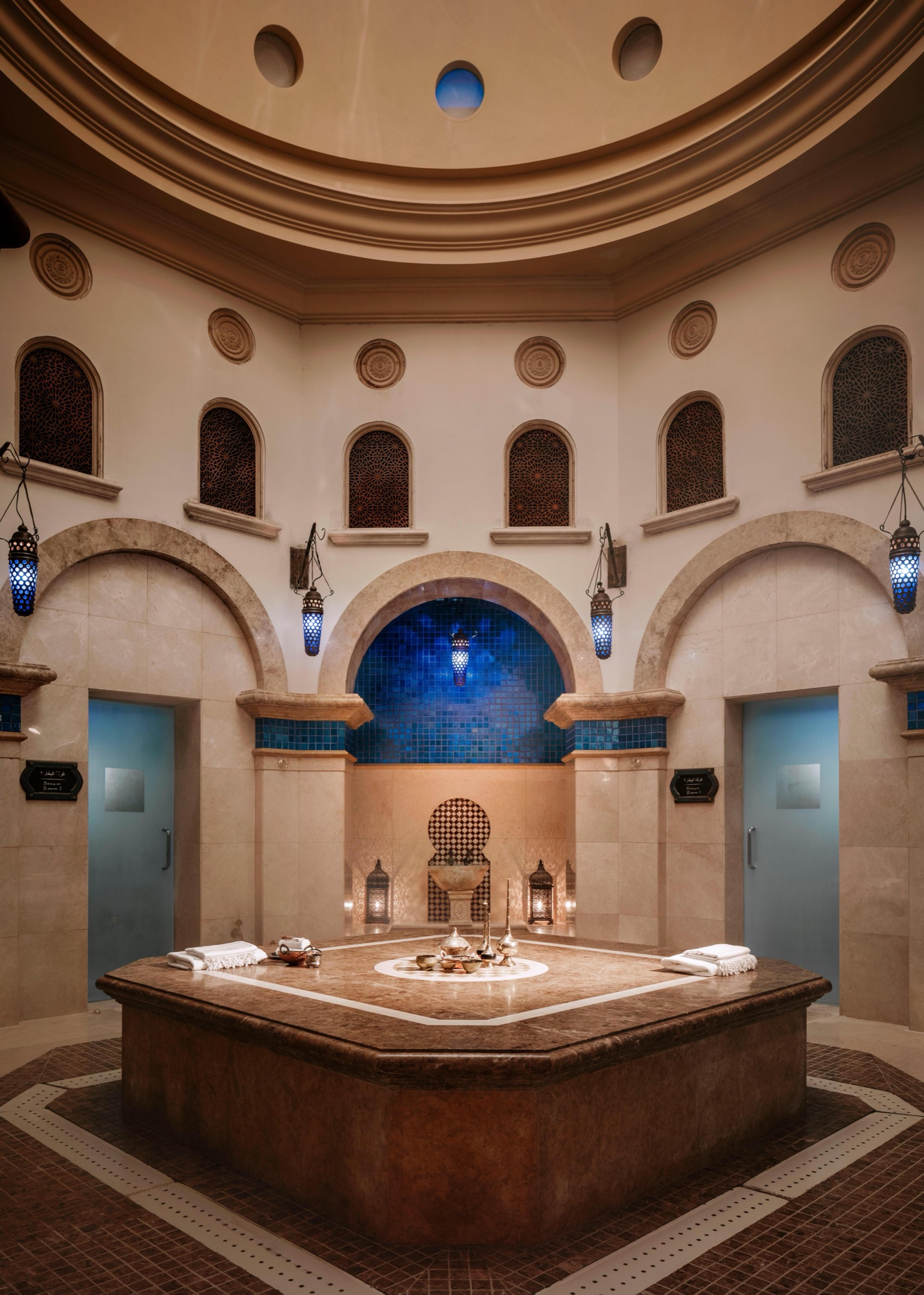 An octagonal stone bench sits at the centre of a domed hammam room with arched blue-tiled niches and hanging lanterns.