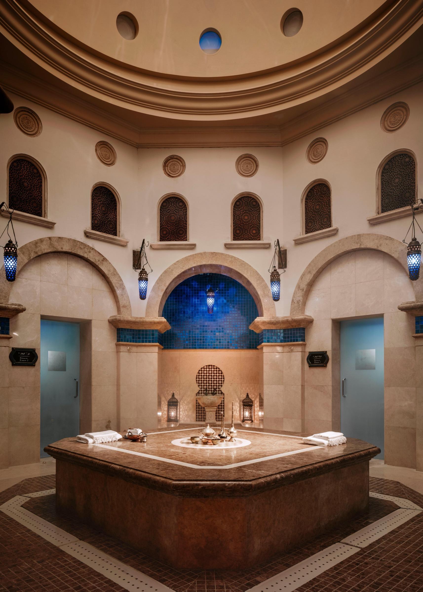 An octagonal stone bench sits at the centre of a domed hammam room with arched blue-tiled niches and hanging lanterns.