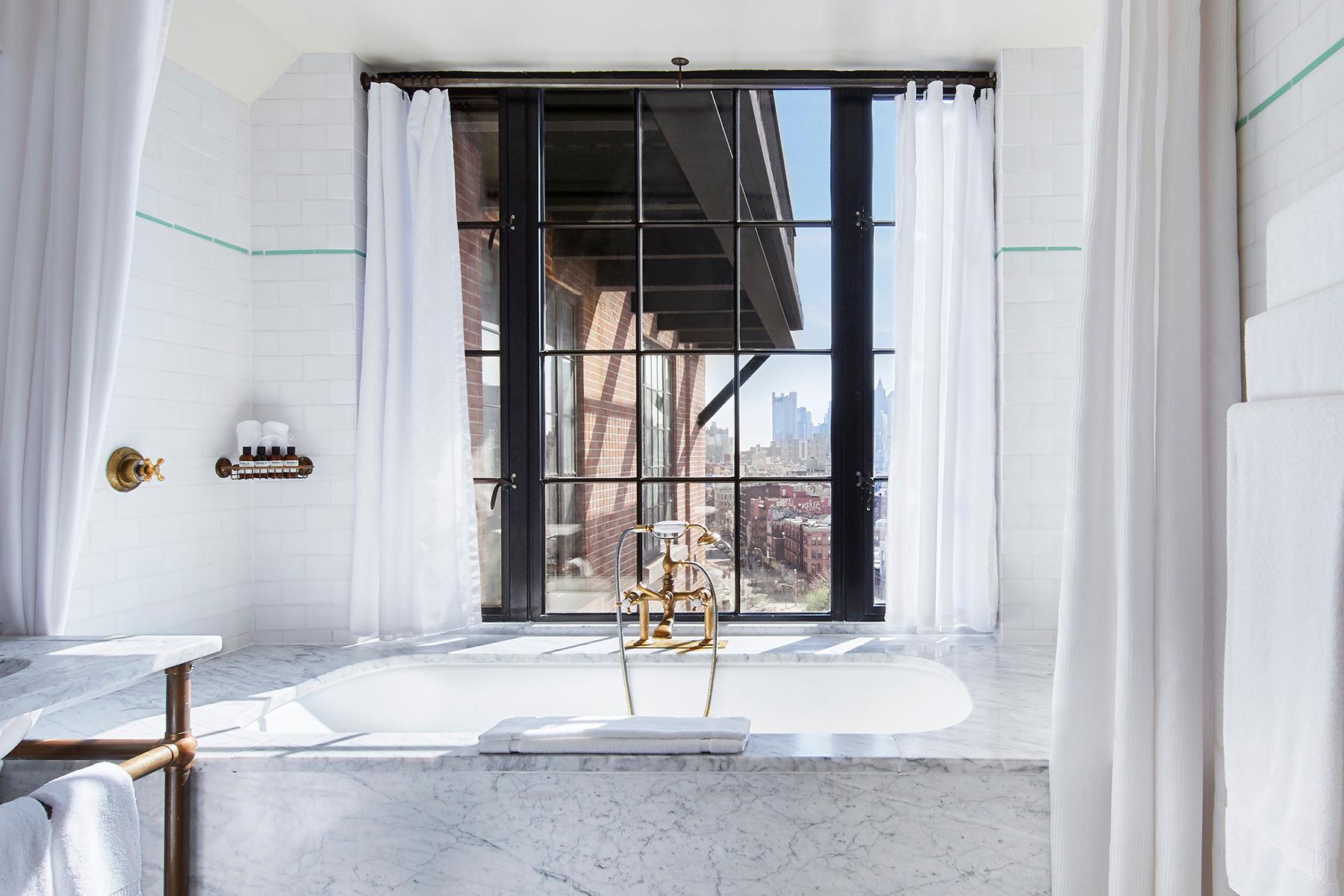 A marble bathroom with a freestanding tub below a tall window looking out over the city, sheer curtains either side.