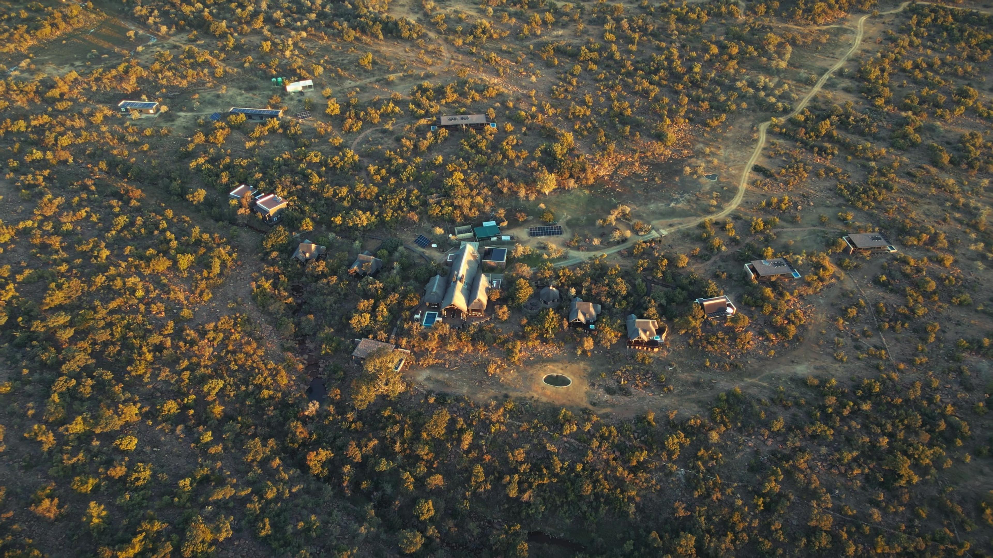 An aerial view of the whole lodge property at golden hour, several thatched buildings linked by paths and scattered across the bush with hills in the distance.